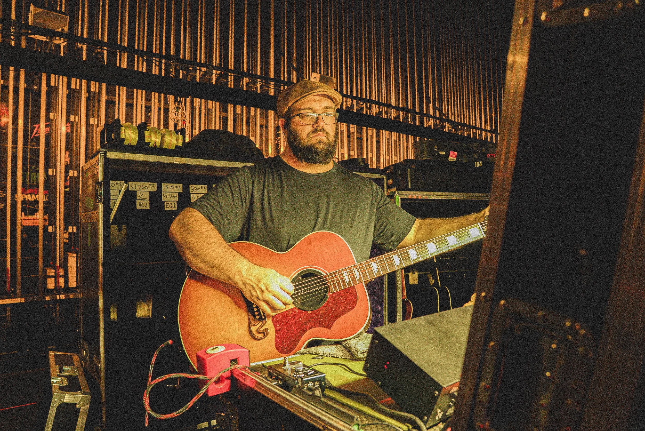 A man playing an acoustic guitar in a music or rehearsal space with equipment and shelves behind him.
