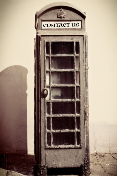Old vintage telephone booth with a sign that says 'CONTACT US' at the top, against a plain wall.