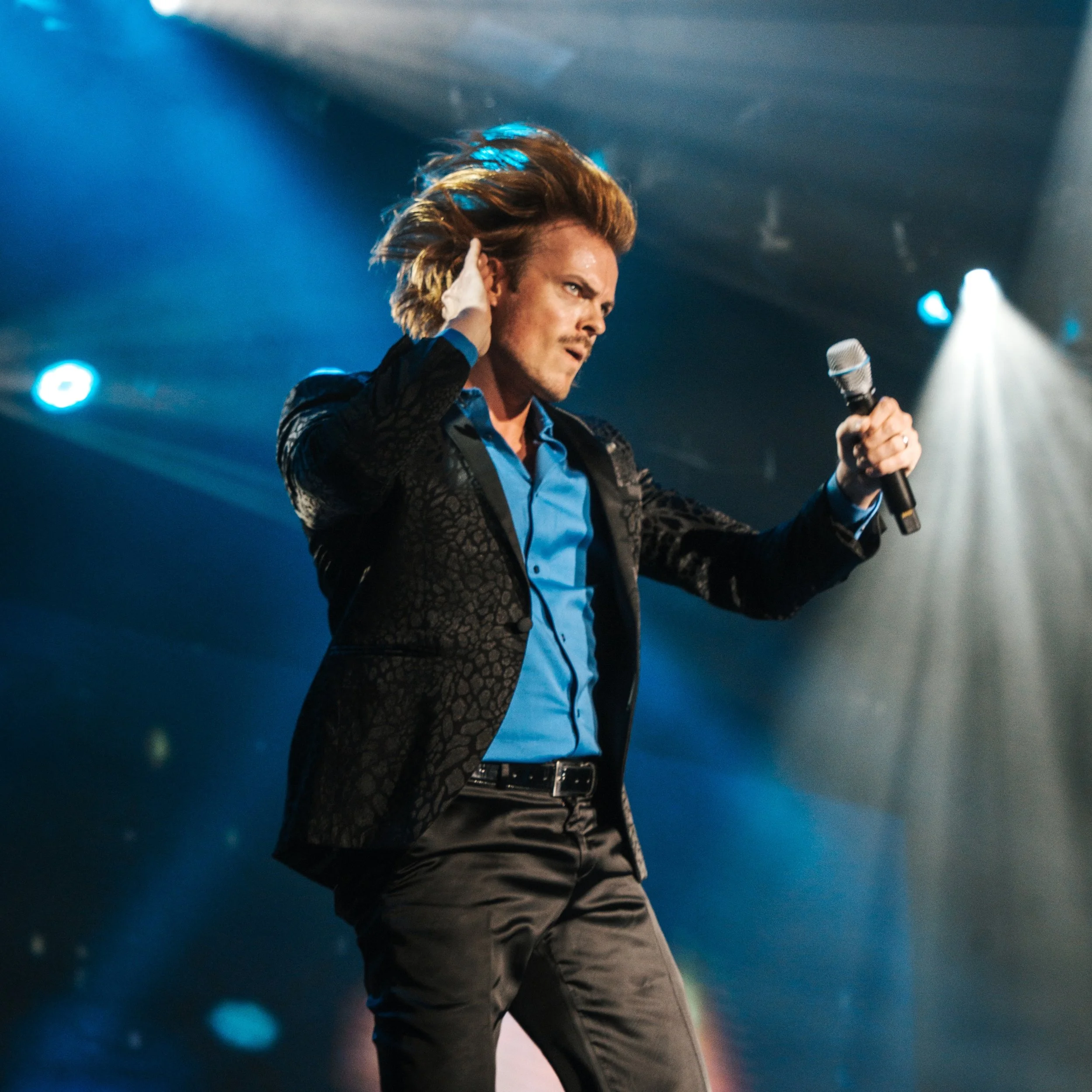 Max Adam, dressed in black suit with blue shirt, with long flowing blond hair as he is in mid jump during a performance holding his hand to his ear to indicate to the audience to sing along.