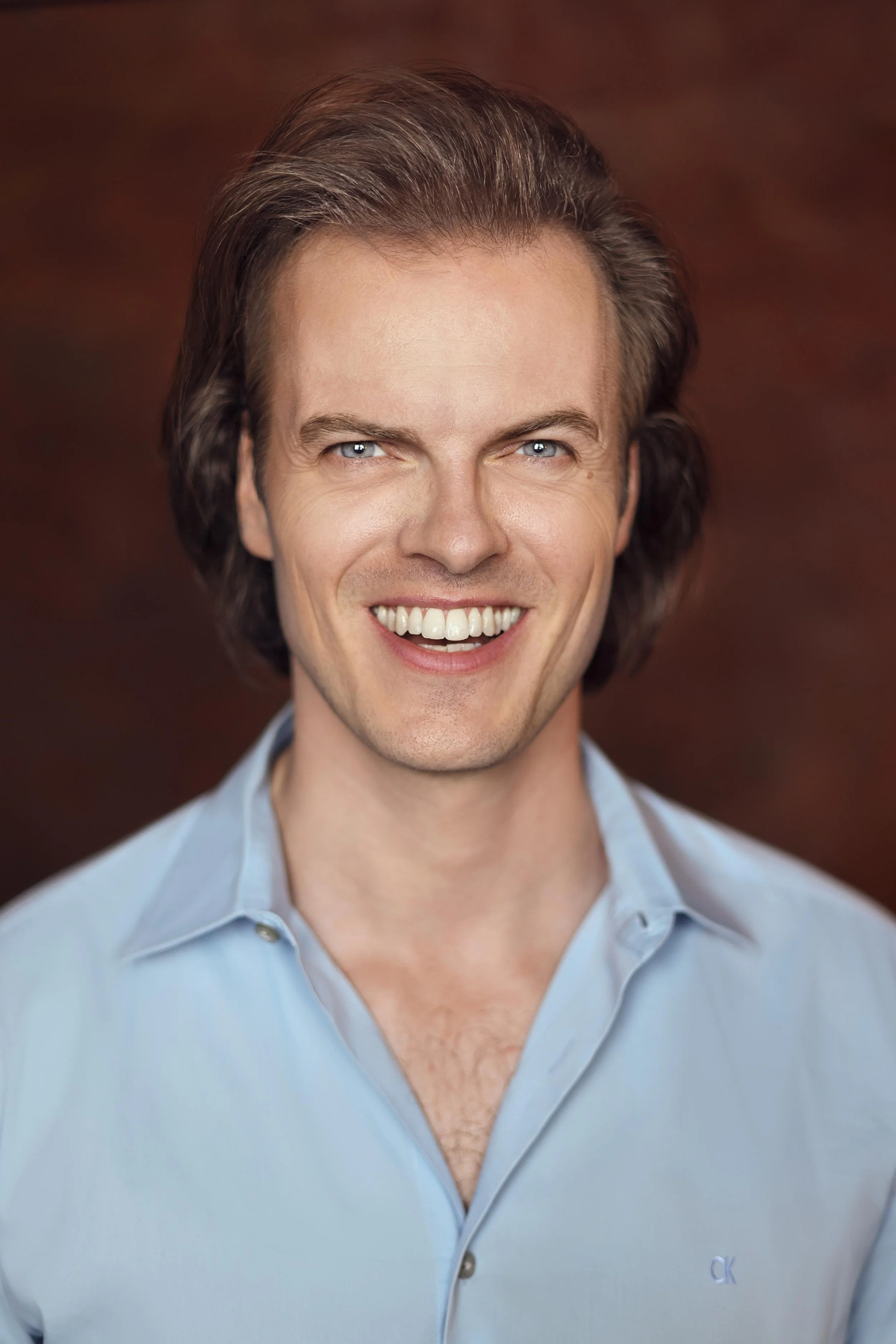 Headshot of a smiling Max Adam wearing a light blue shirt against a dark brown background, studio portrait.