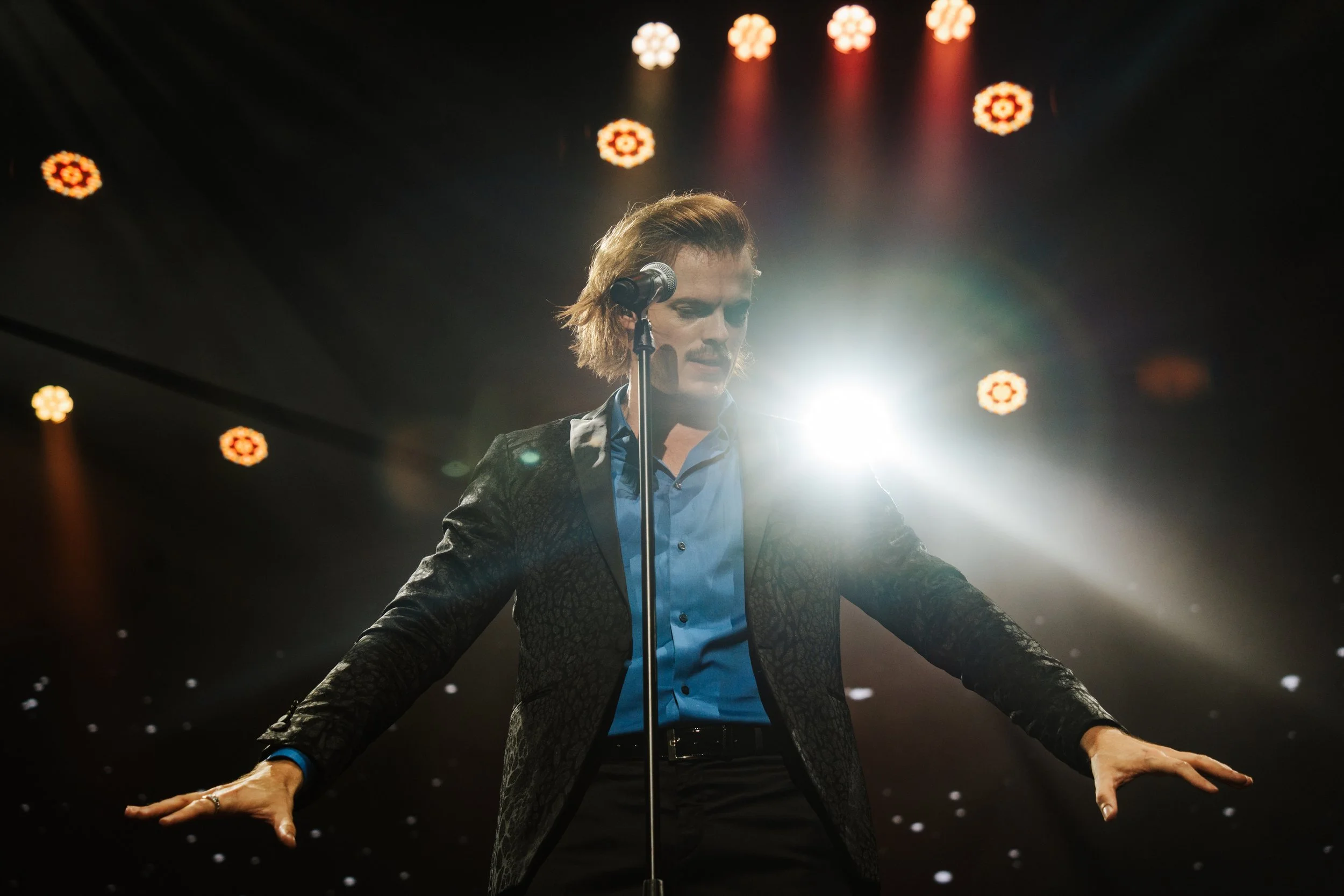 A backlit Max Adam on a stage, wearing a black suit with blue shirt, standing in front of a microphone in a serious performance pose with arms extended.