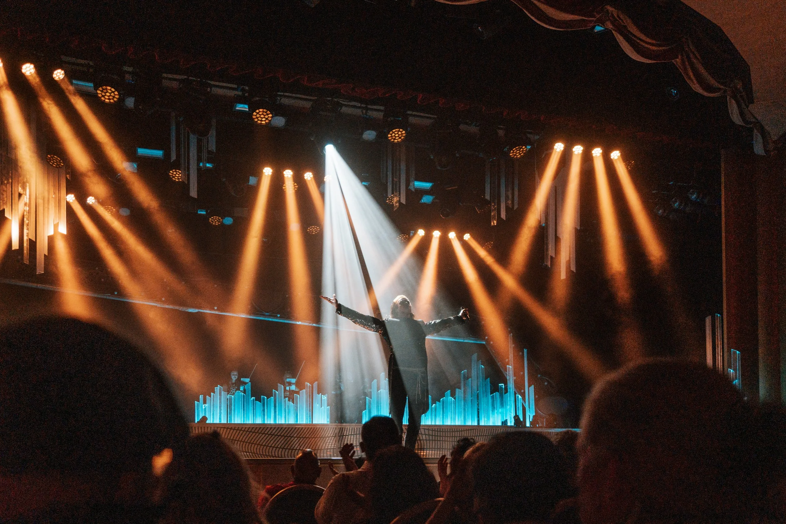 Max Adam stands on stage with arms raised, illuminated by multiple bright stage lights, with an applauding audience in the foreground in full ovation
