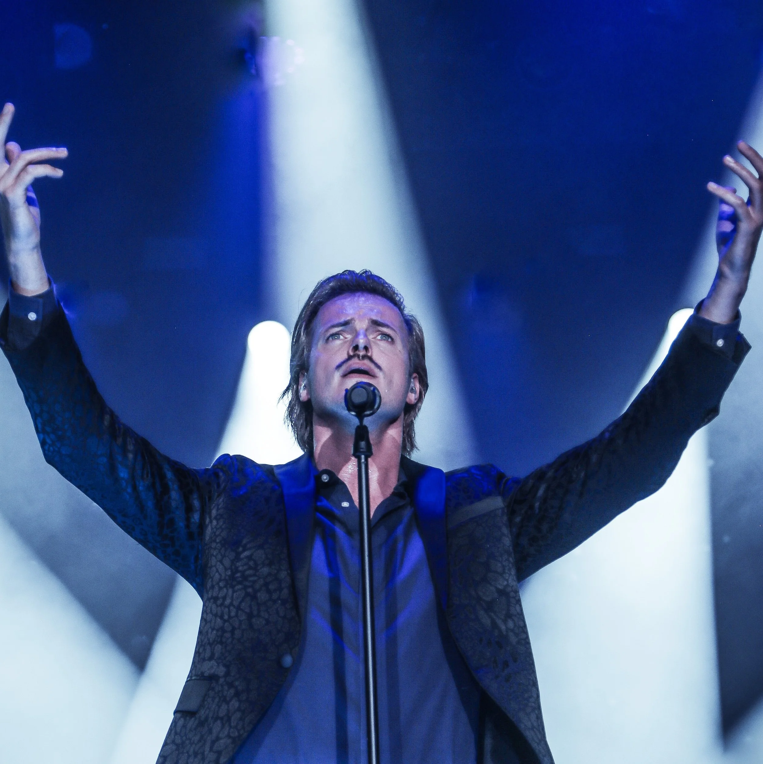 Max Adam performing Nessun Dorma, standing in front of a microphone, looking upward with an expression of awe and prayer. The background features blue and white lights creating a dramatic atmosphere.