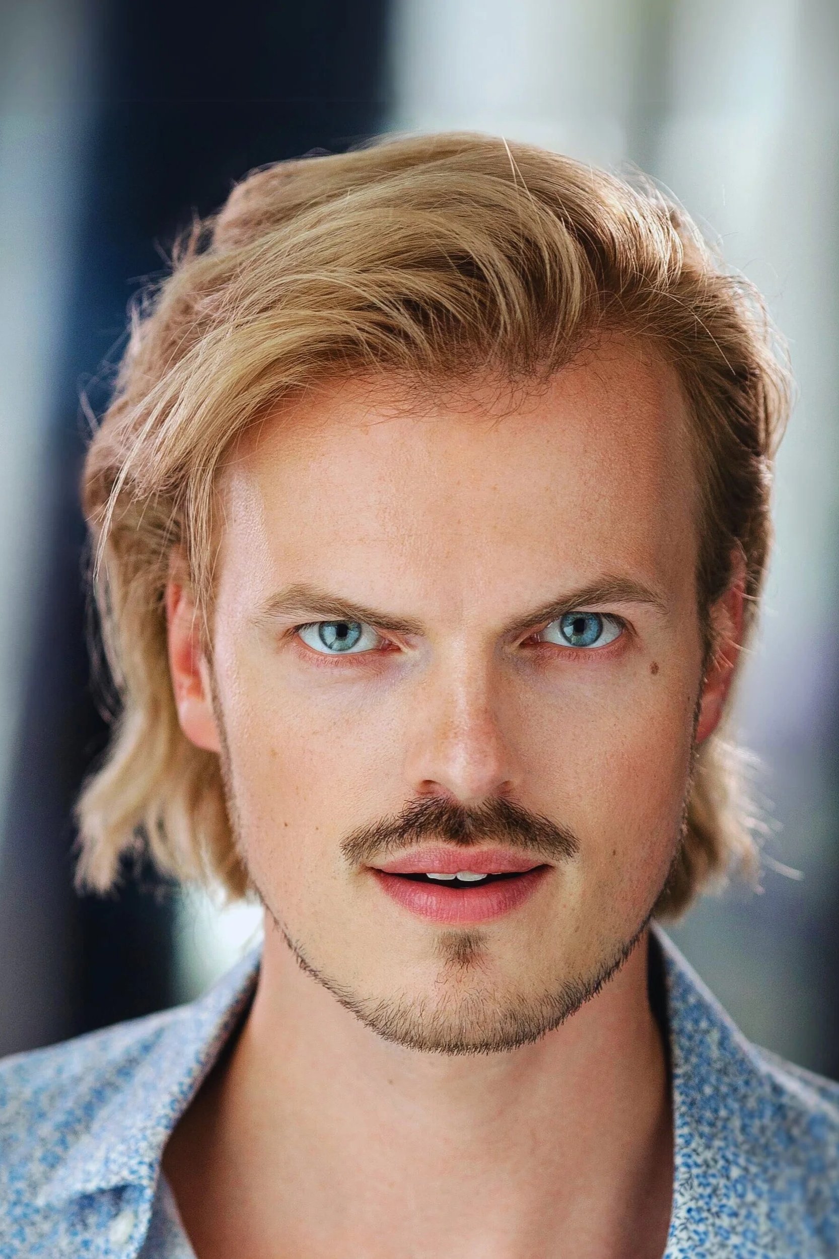 Close-up Headshot of Max Adam with sun bleached hair wearing a light blue shirt against a blurred blue background, studio portrait.