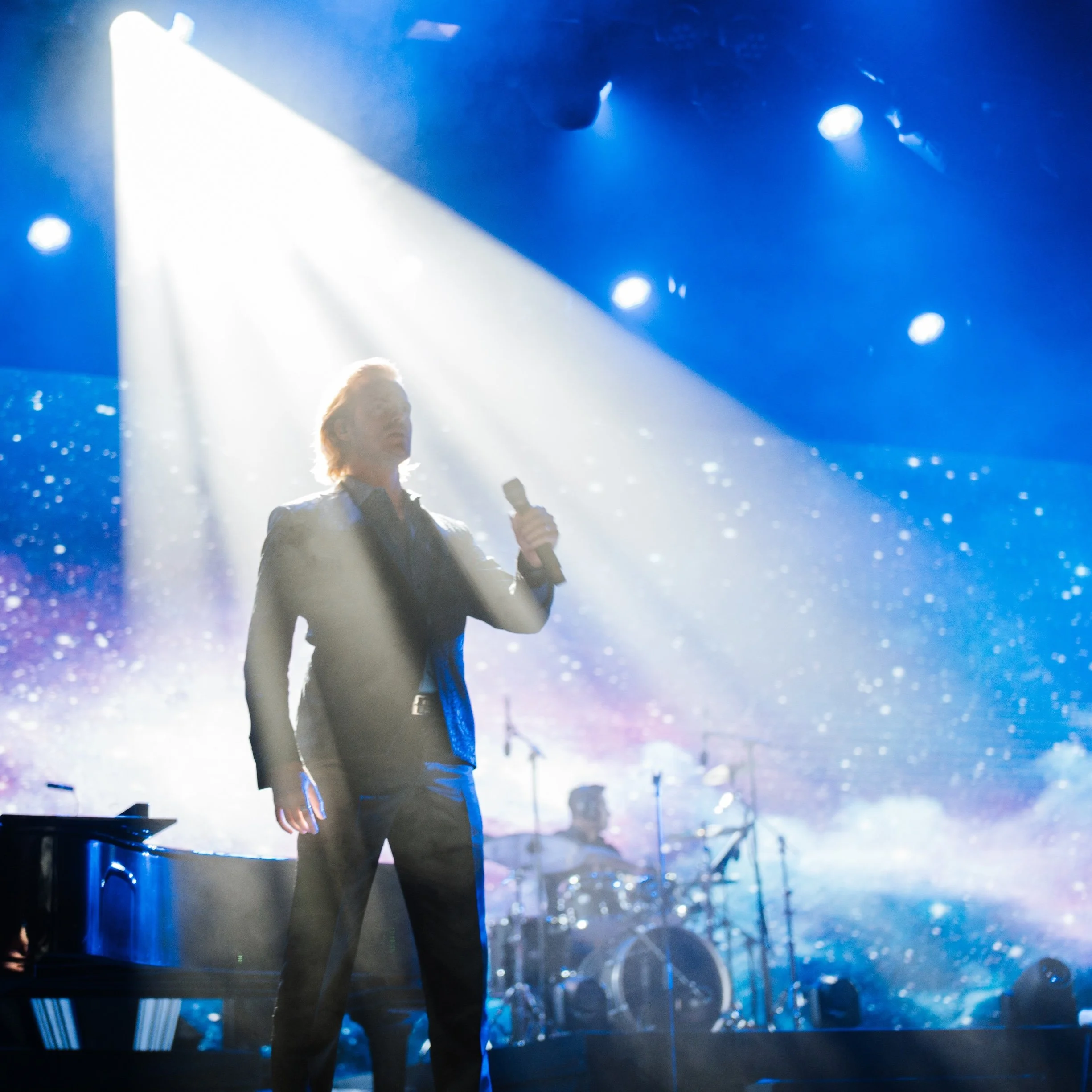 Max Adam holding a microphone on stage with blue galaxy background and white lighting effects.