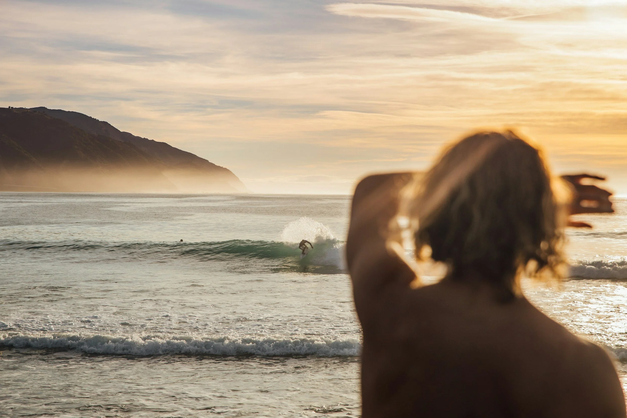 A woman with curly hair is standing on the beach, watching a surfer ride a wave during sunset with mountains in the background.