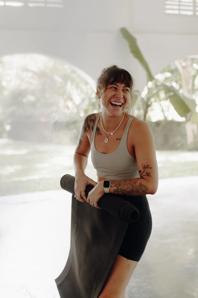 Tattooed EarthLinks guest holding a rolled yoga mat, smiling indoors in natural light.