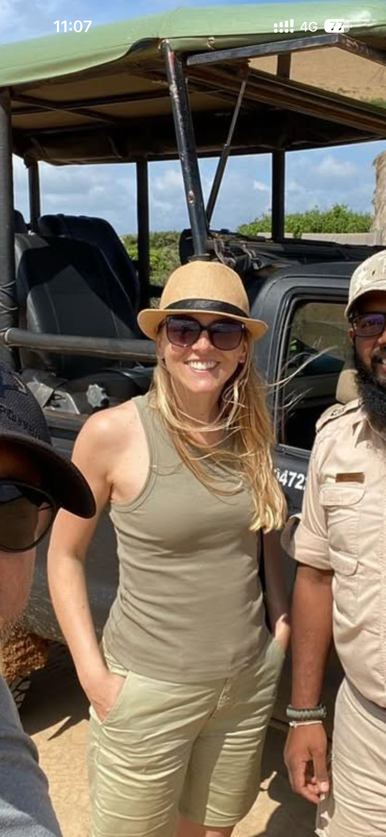 A woman wearing a wide-brimmed hat and sunglasses, smiling while standing next to a man in safari attire, with a green open-sided vehicle and lush greenery in the background.