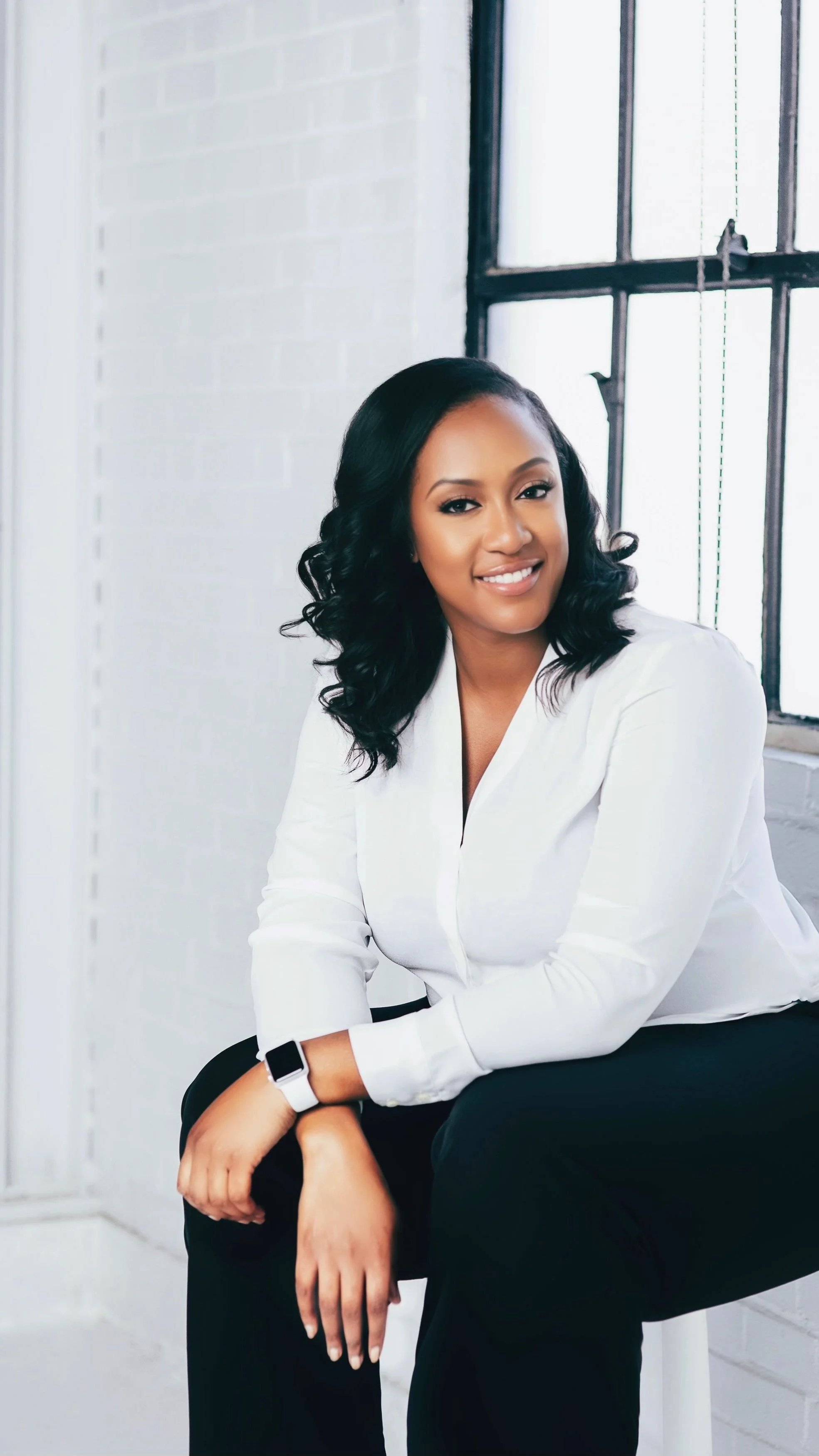 A woman with black wavy hair smiling, wearing a white blouse and black pants, sitting near a large window in a bright room.