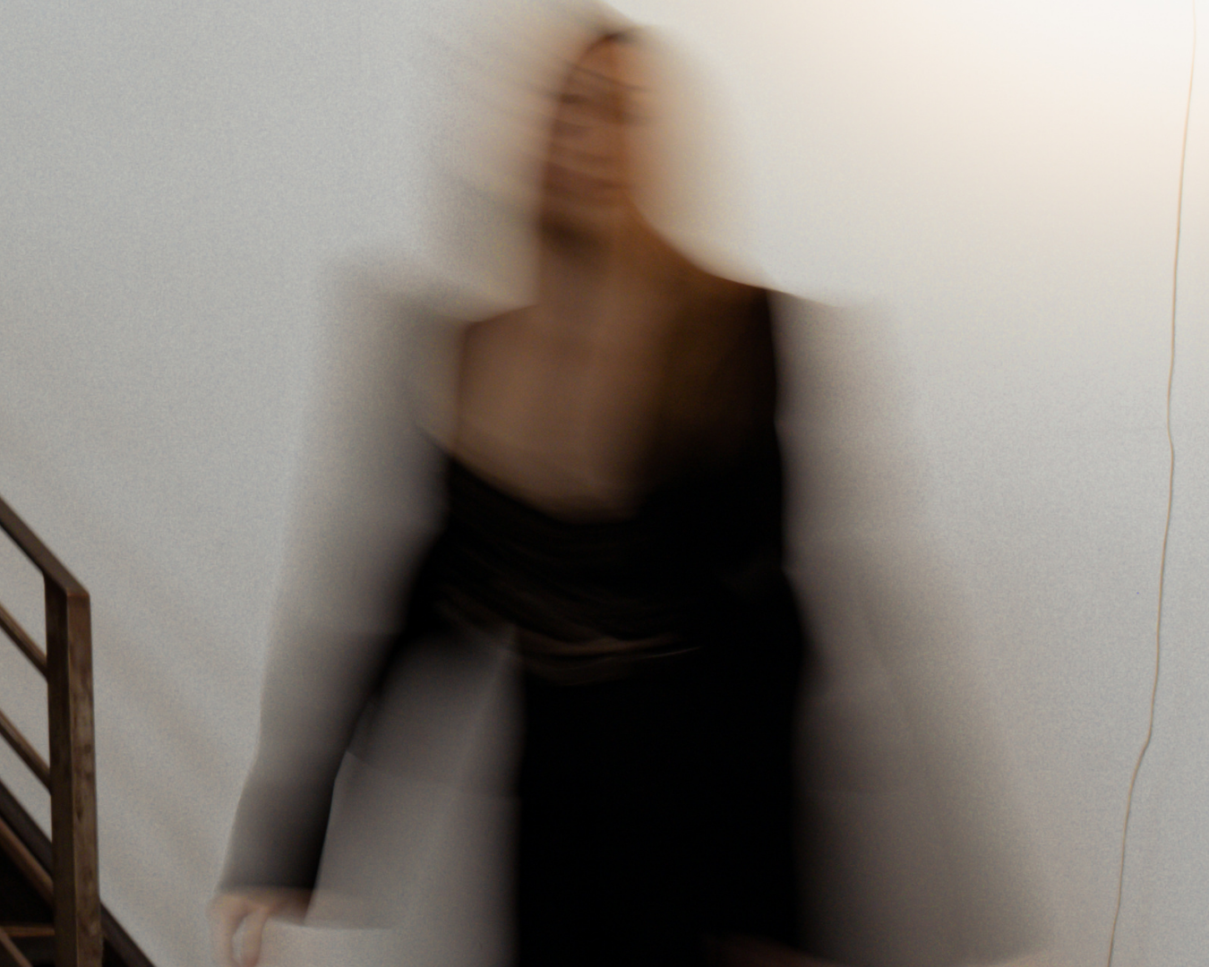 Blurred image of a woman standing against a plain white wall, with a wooden staircase visible on the left side.