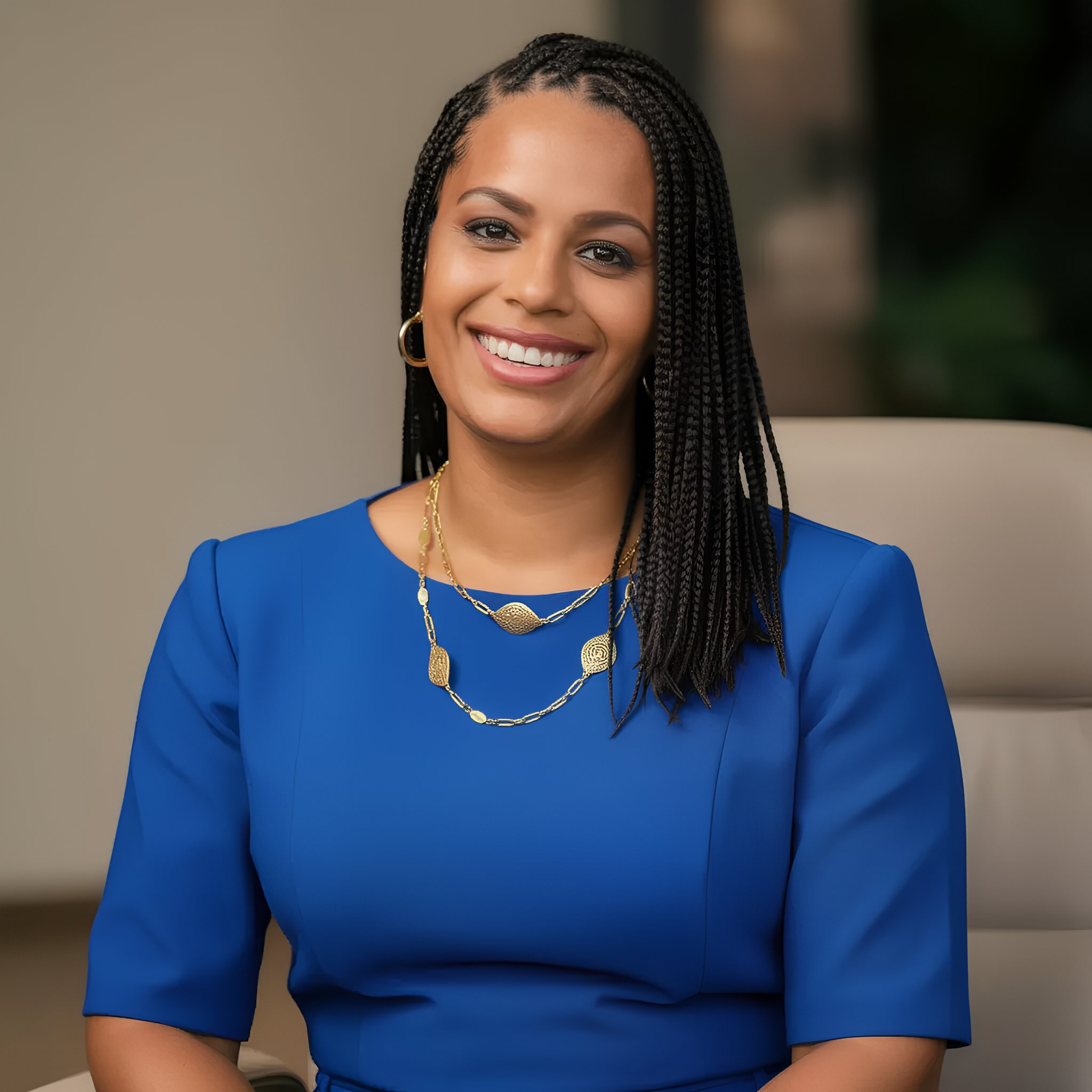 A smiling African American woman with long braided hair wearing a blue dress and gold jewelry, sitting on a beige chair indoors.