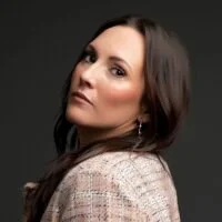 A woman with long dark hair looking over her shoulder against a dark gray background.