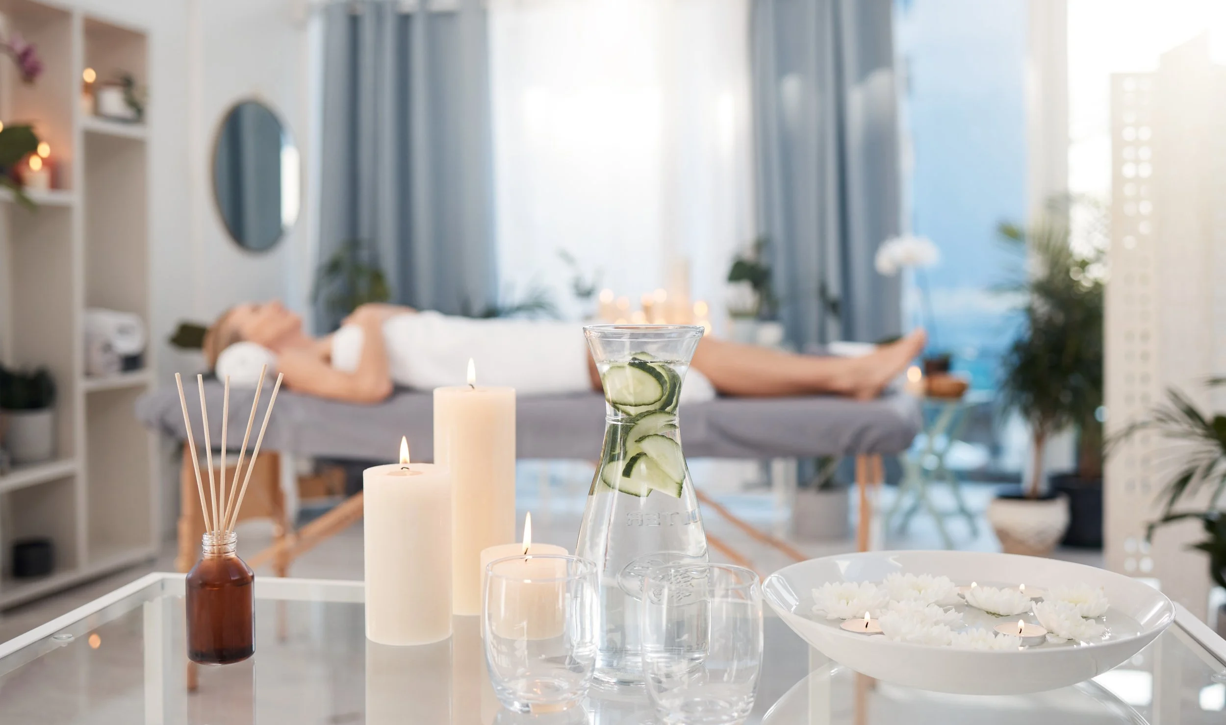 Spa or massage room with a woman lying on a massage table in the background, and candles, a glass pitcher with cucumber slices, and floating candles on a table in the foreground.