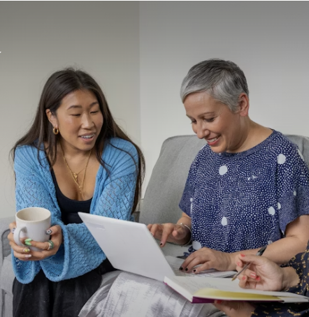Two women sitting on a couch looking at a tablet, one holding a mug, smiling and engaging in conversation.