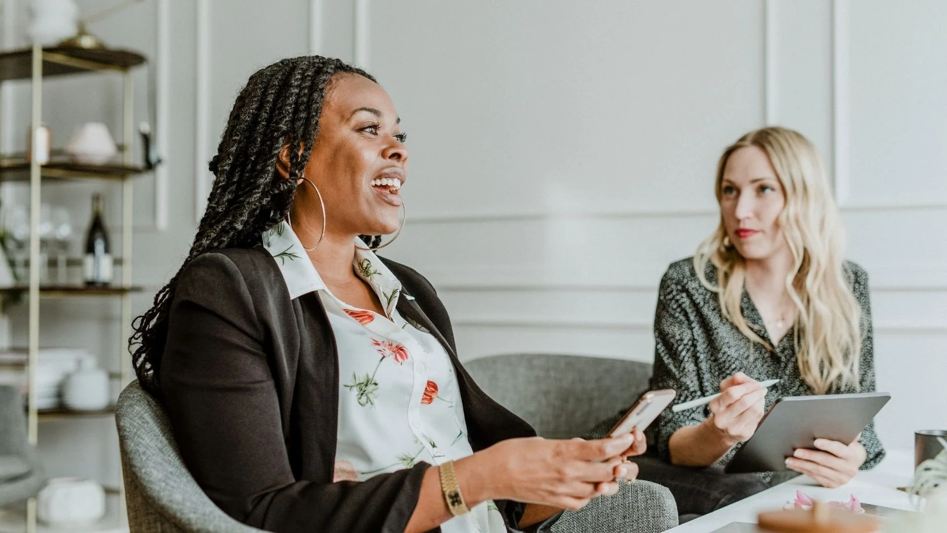 Two women having a discussion at a meeting, one holding a smartphone and the other a tablet, in a modern office setting.