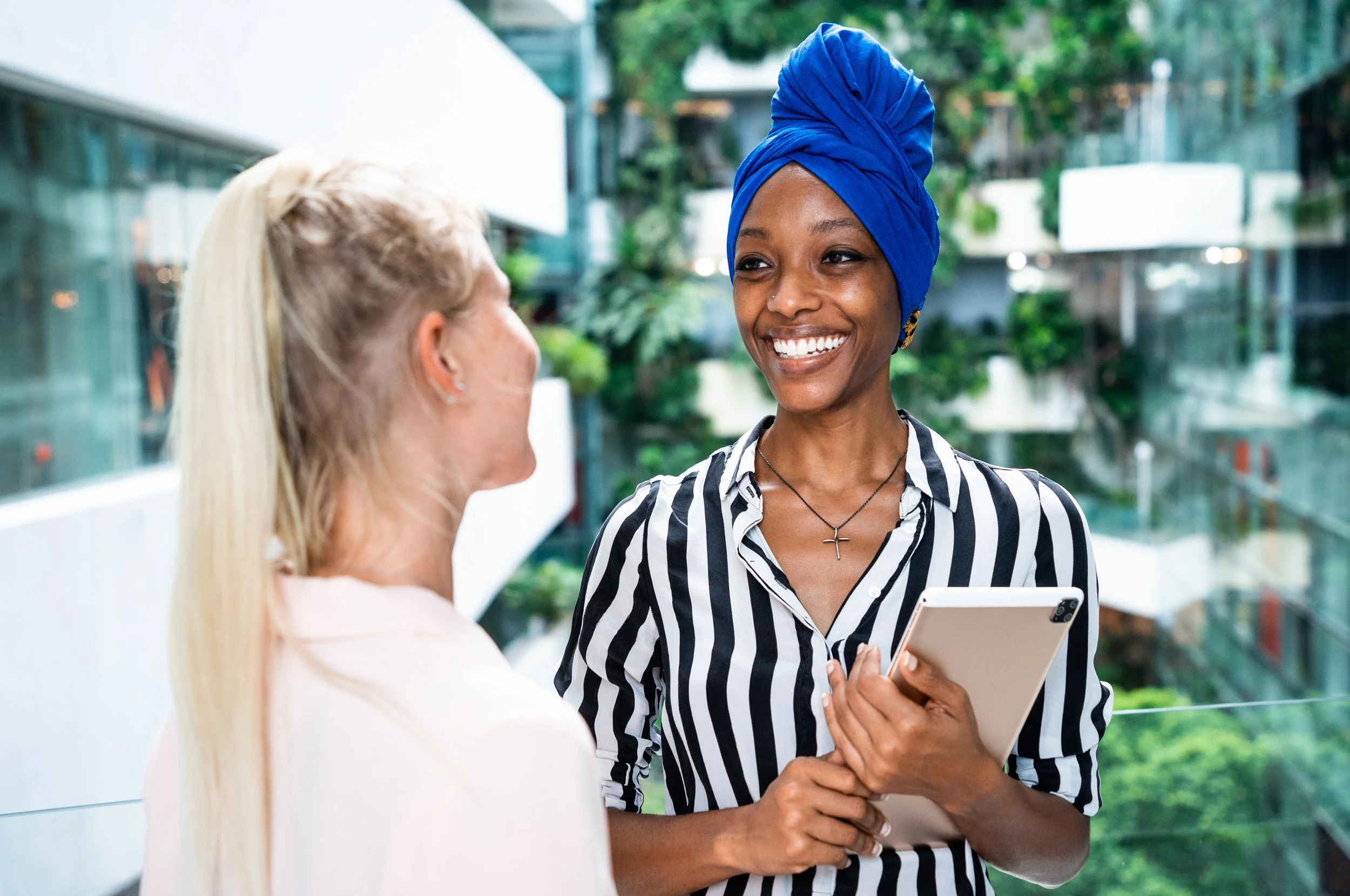 Two women conversing: one with long blonde hair, seen from behind, and the other with dark skin wearing a blue headwrap, striped shirt, and holding a tablet, smiling outdoors in an urban setting with glass buildings and greenery.