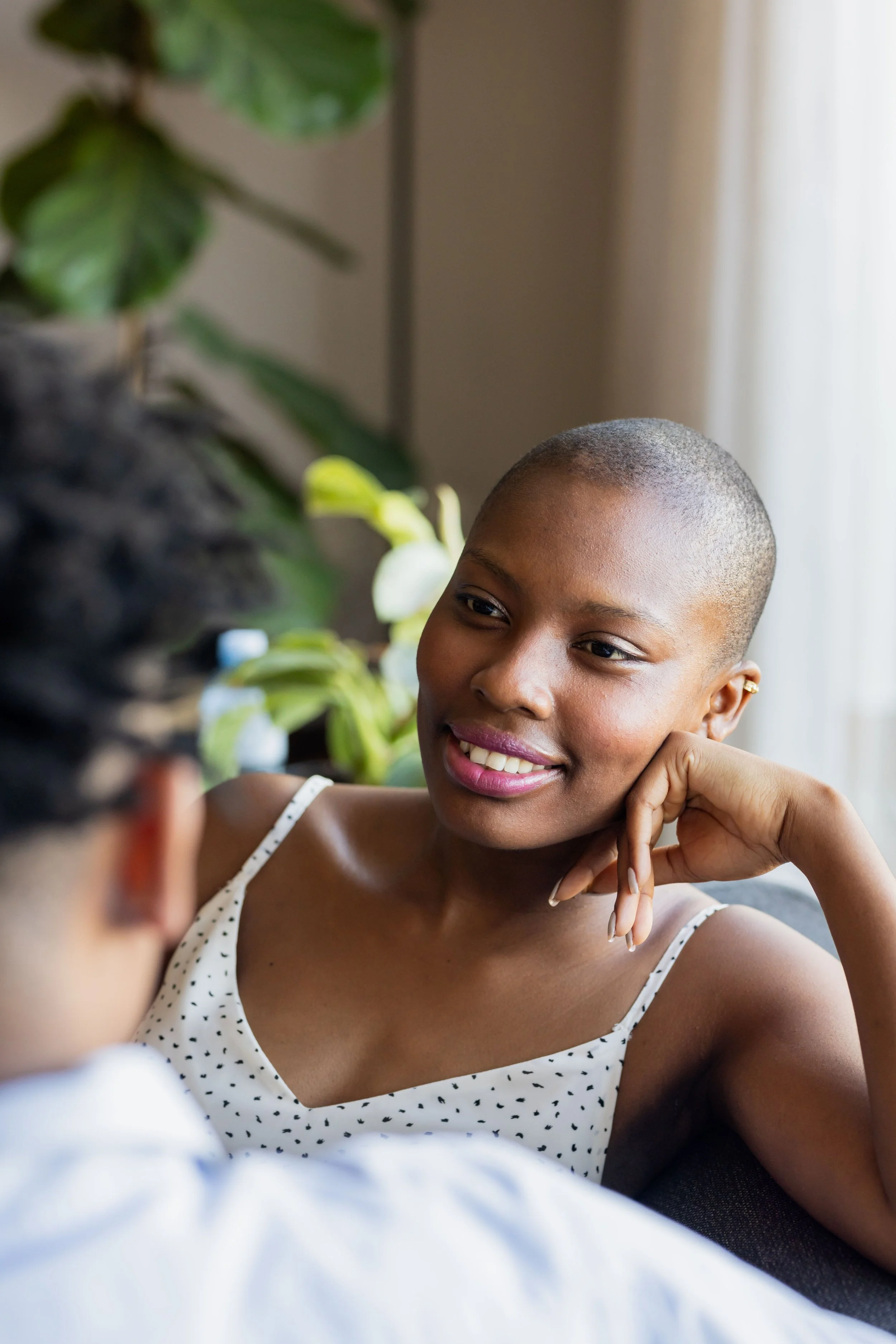 A woman with a short hairstyle, wearing a white polka dot top, smiling and listening to a person with glasses, indoors near a window with natural light and green plants in the background.