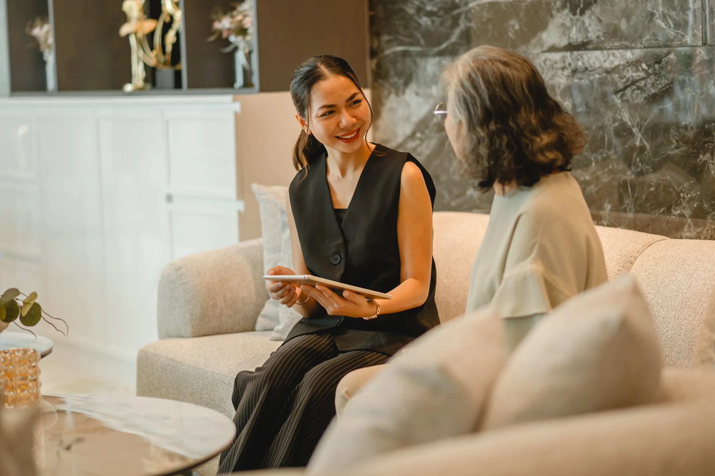 Two women sitting on a beige sofa, engaged in conversation, one holding a tablet, in a modern, elegantly decorated room.