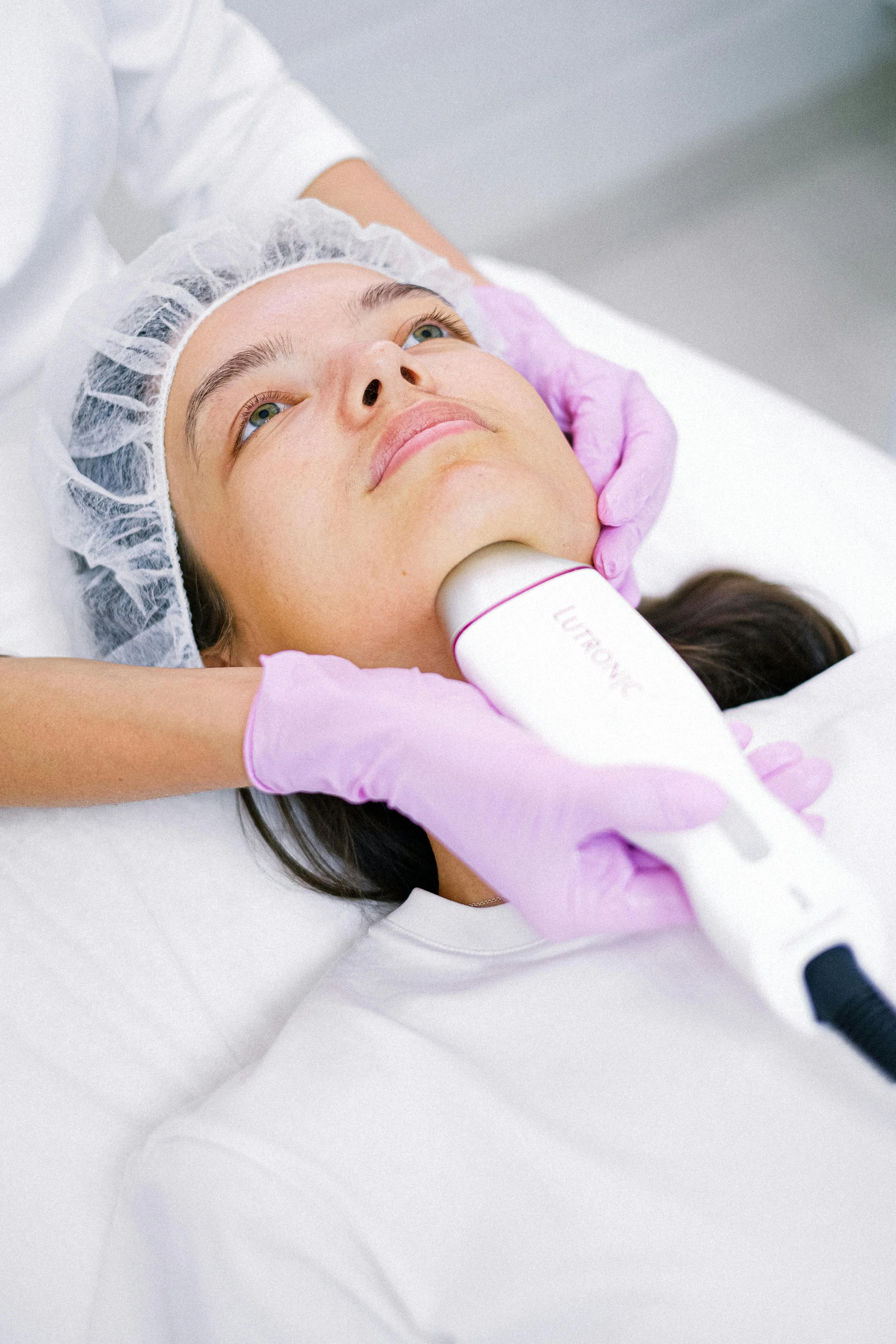 A woman lying on a treatment bed with a head cap, receiving a facial treatment with a device labeled "LUMIONE" by a professional wearing pink gloves.