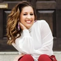 A woman smiling and sitting outdoors, wearing a white top and red pants.