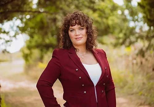 A woman with curly brown hair wearing a maroon blazer and white top standing outdoors with trees in the background.