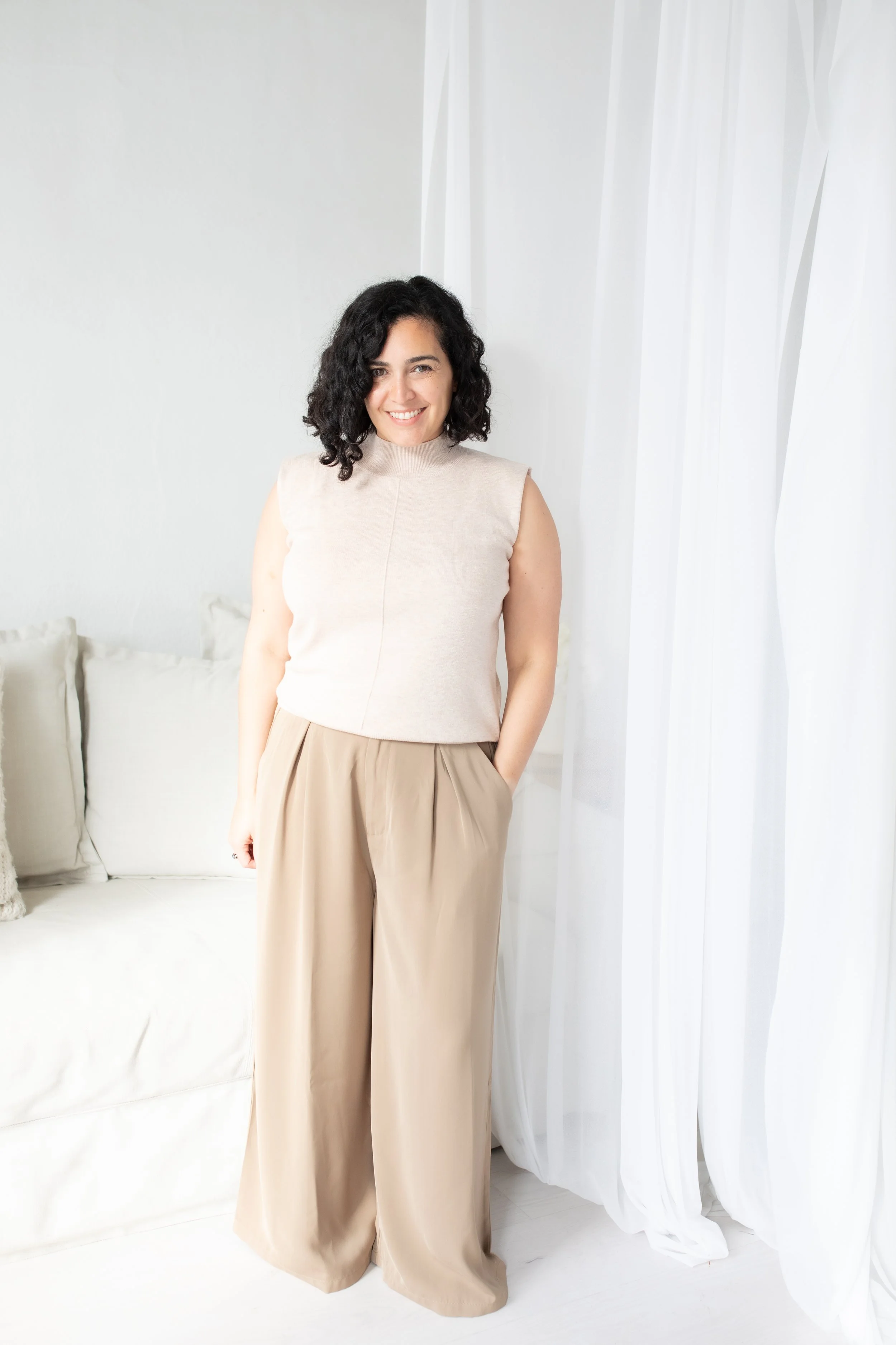 A woman with curly black hair smiling, wearing a beige sleeveless top and wide beige pants, standing in a bright room next to white curtains.