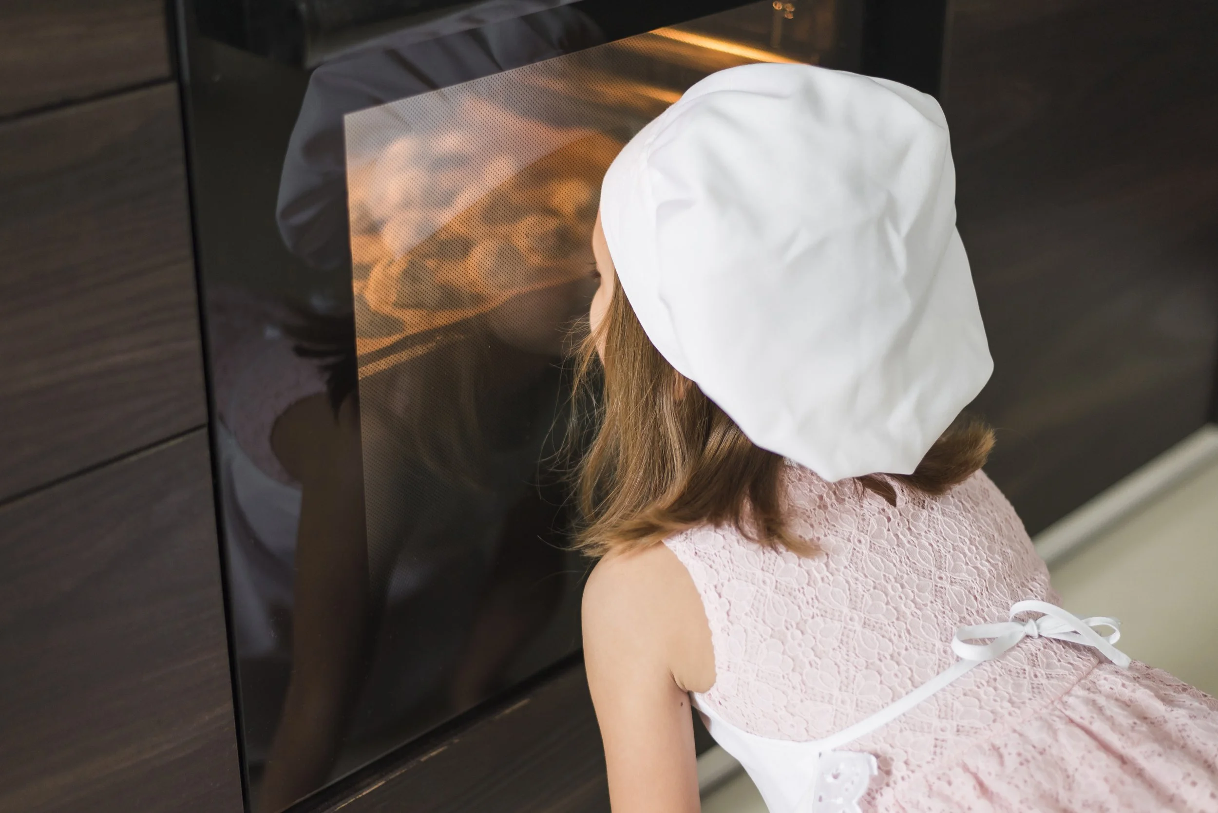 A young girl wearing a white chef's hat and pink lace dress watches baked goods inside an oven.