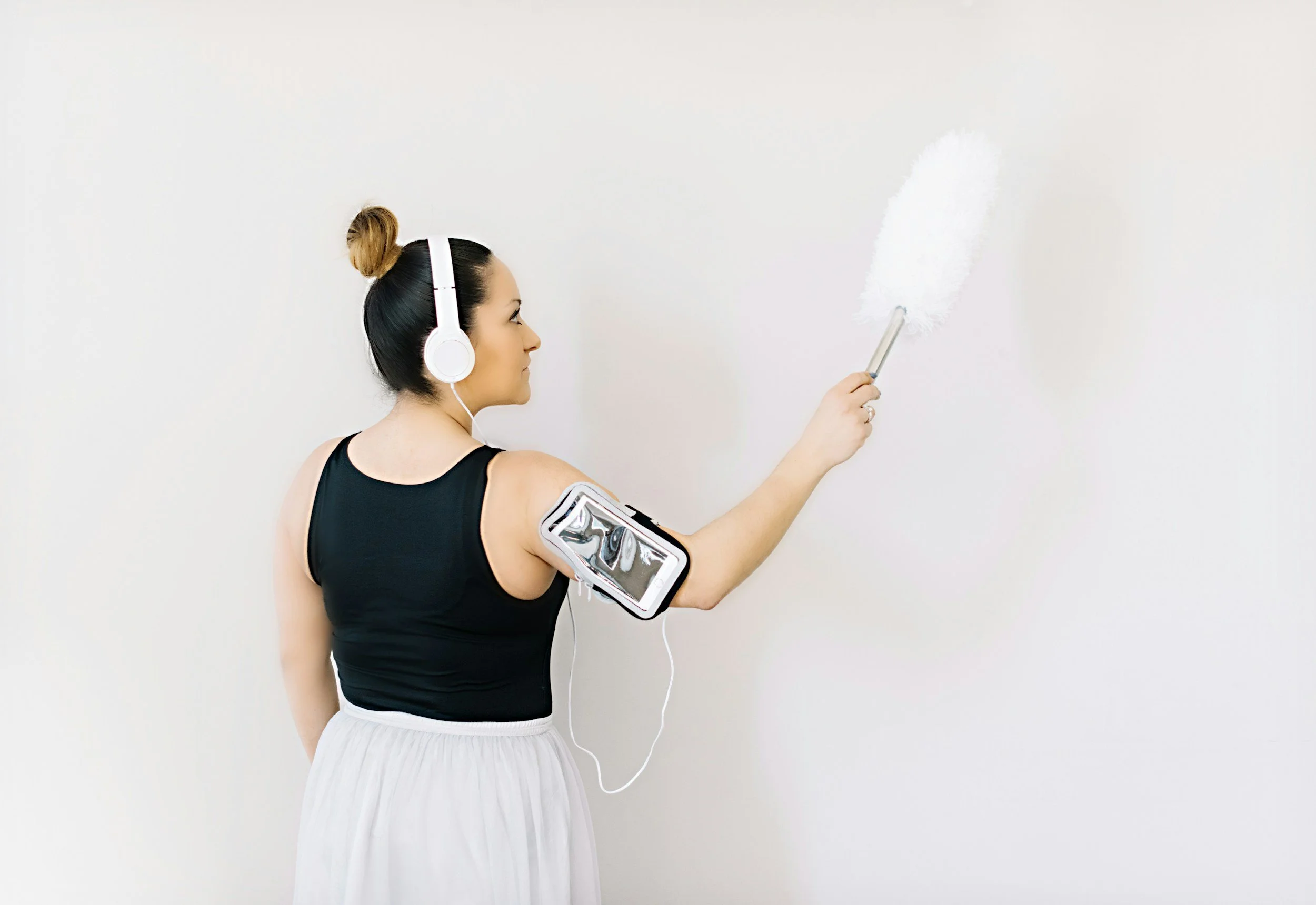 Woman in black sleeveless top and white skirt cleaning a white wall with a duster, wearing headphones and with a smartphone in a sports armband.