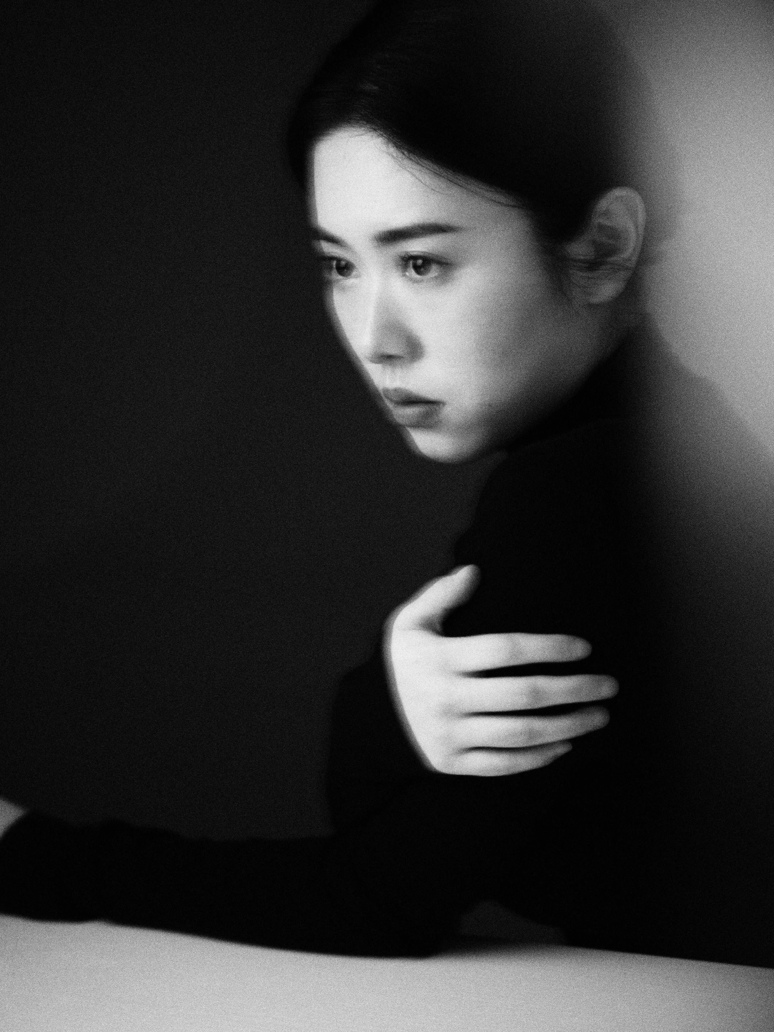 Black and white portrait of a young woman with dark hair, gazing thoughtfully to the side, with one hand resting on her shoulder, wearing a dark long-sleeved top.