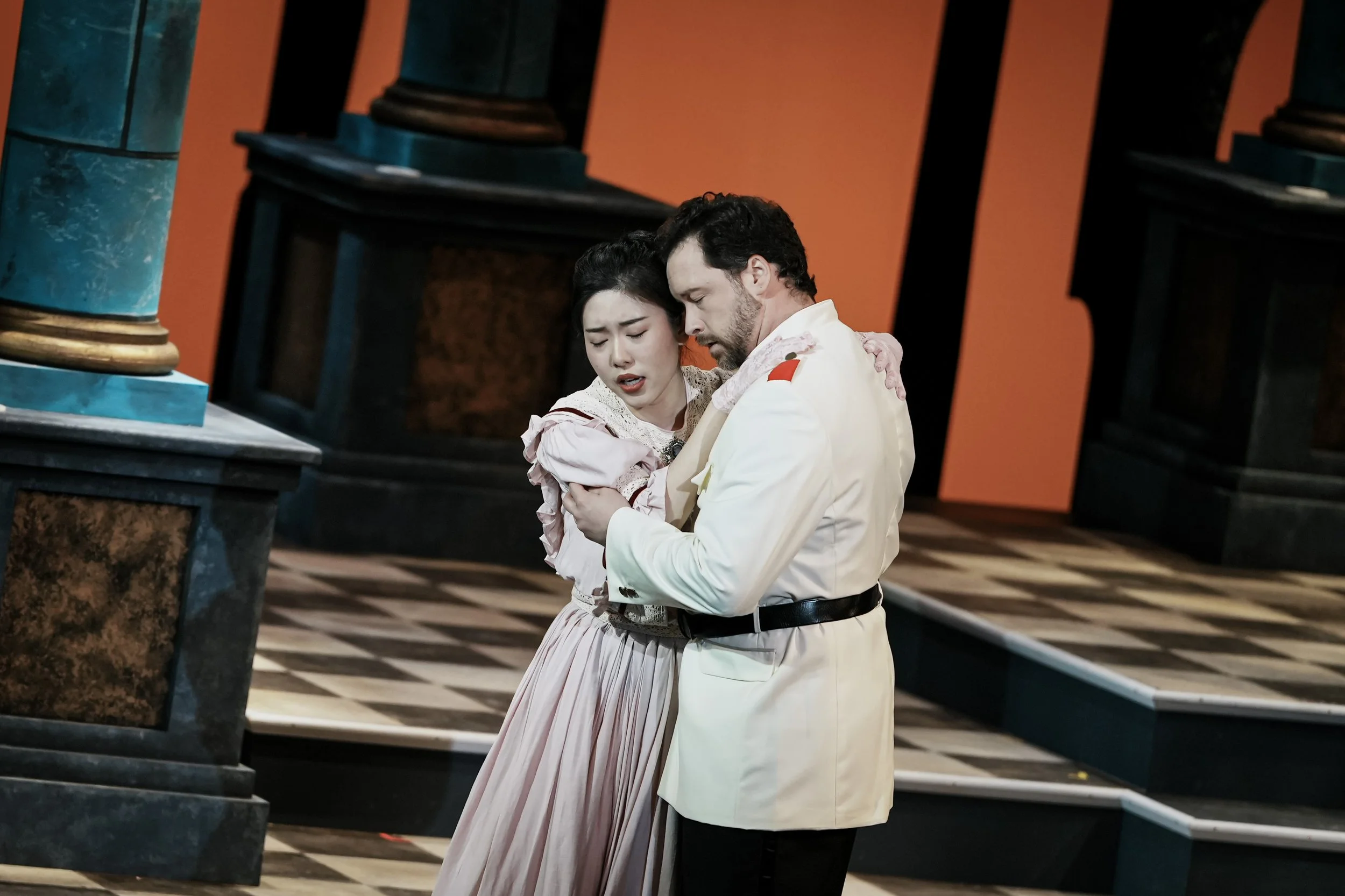 A man and woman embrace passionately on stage in a theatrical production; the woman appears distressed, the man is dressed in a white military uniform with a red epaulet; stage with black and brown pillars and a checkered floor in the background.