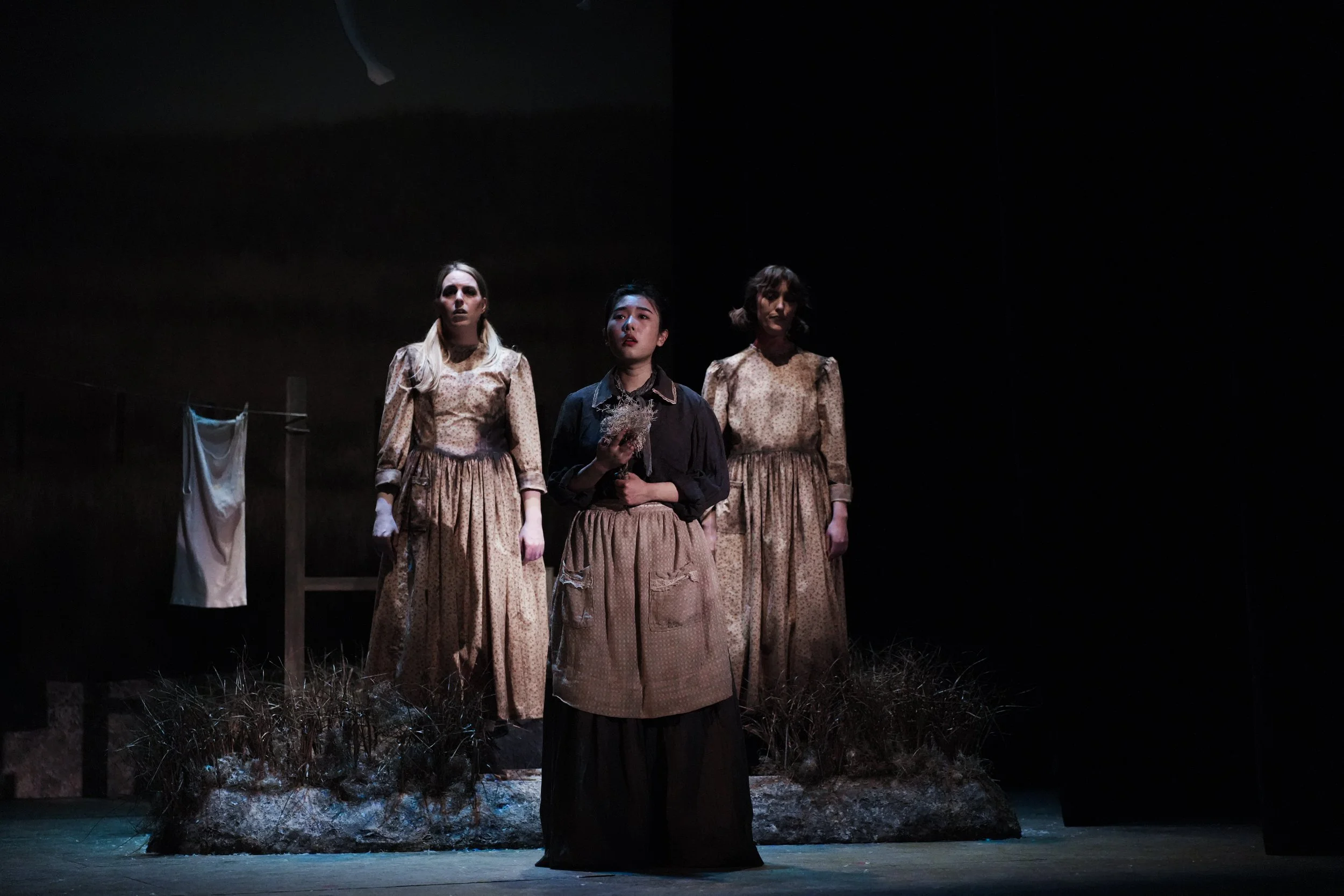 Three women standing on a stage, with one holding a bouquet, in a dark theater setting.