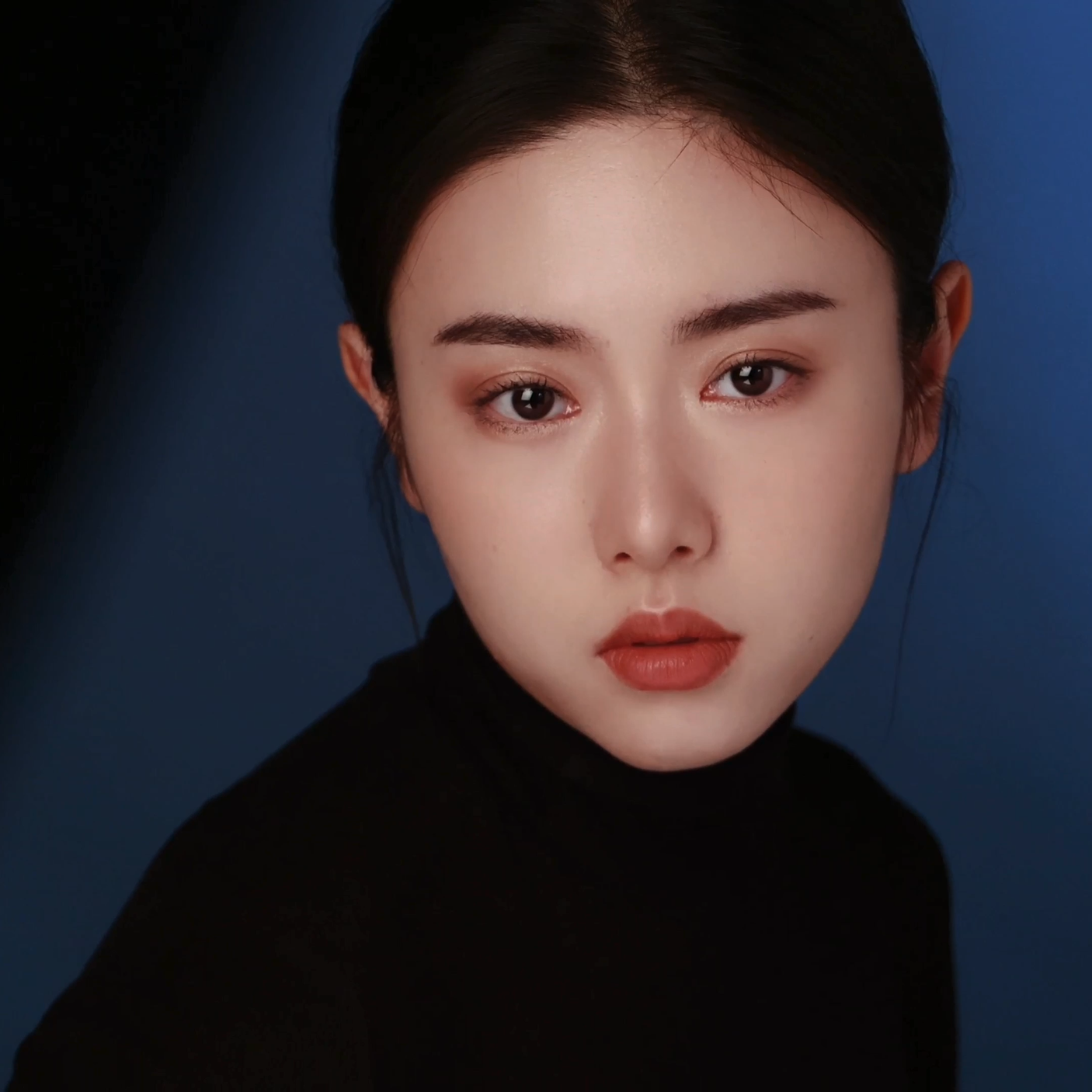 Close-up portrait of a young woman with dark hair, wearing a black turtleneck, against a dark blue background.