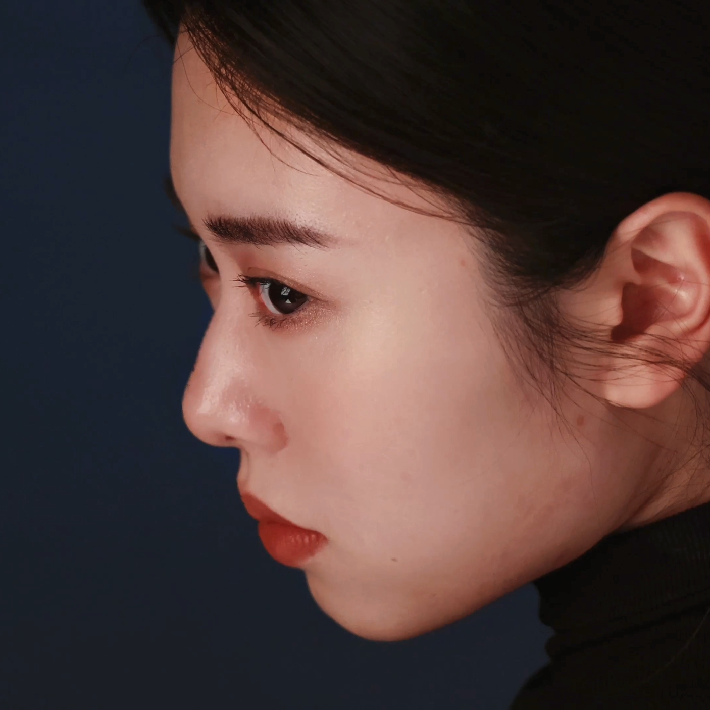 Close-up of a woman's side profile with smooth skin, dark hair, groomed eyebrows, and red lipstick, set against a dark background.