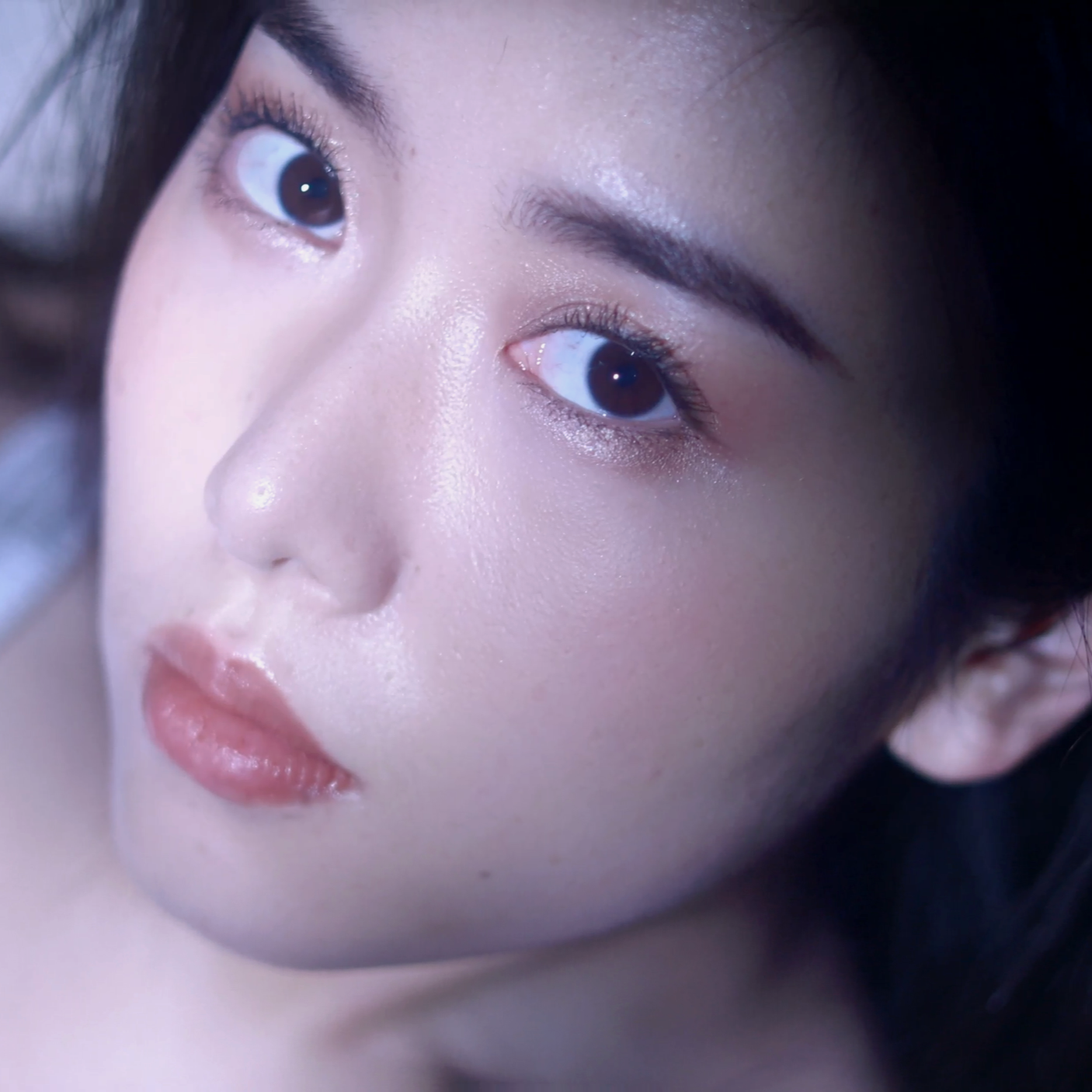 Close-up of a woman's face with perfect skin, dark eyes, and pink lips, looking slightly to the side.