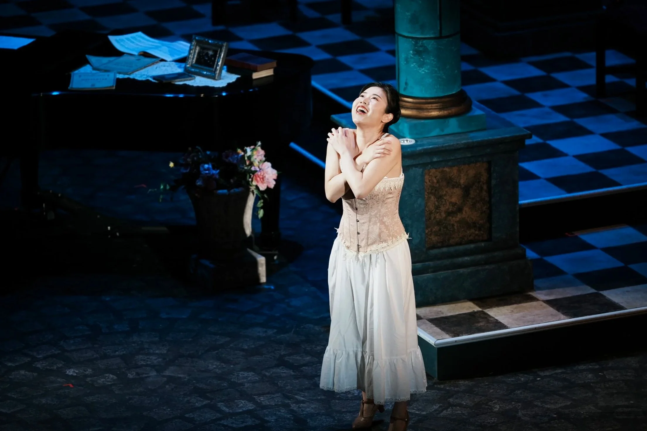 A woman on stage dressed in vintage clothing, standing with arms crossed over her chest, smiling with eyes closed in a theatrical setting.