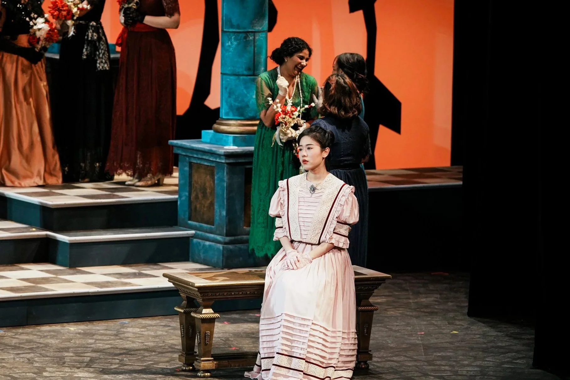 A stage scene from a theatrical play with women in vintage costumes; one woman in a pink dress is seated in the foreground, while others stand and converse in the background against an orange backdrop.