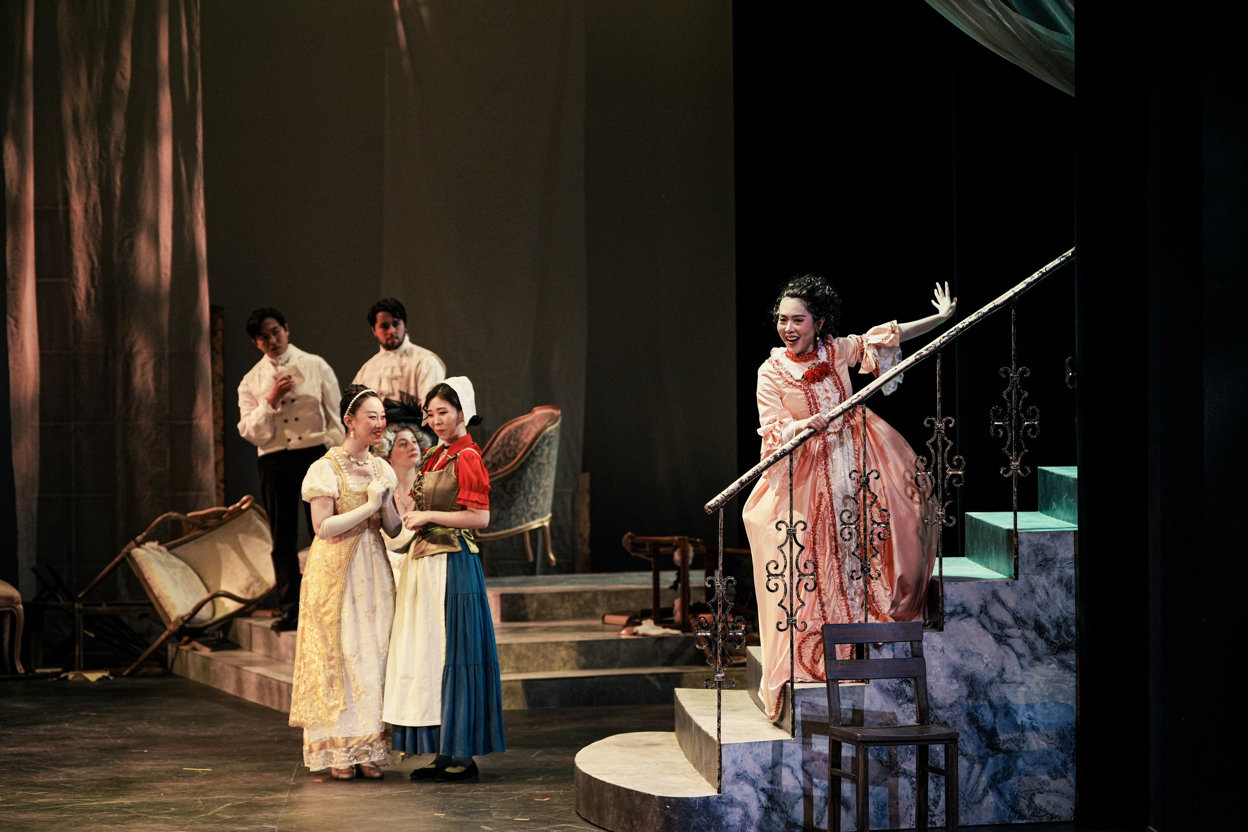Theater scene with performers on stage: two women in elaborate dresses in the foreground, one gesturing with her hand on stairs, three people in the background with two men and one woman, and a set with furniture and props.