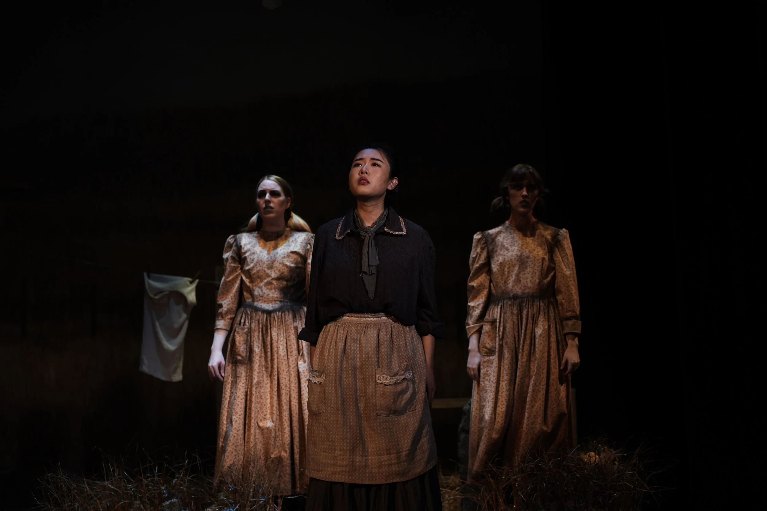 Three women standing on a dark stage, one in front wearing a black blouse and an apron, with two women behind wearing long patterned dresses, in a theatrical performance setting.