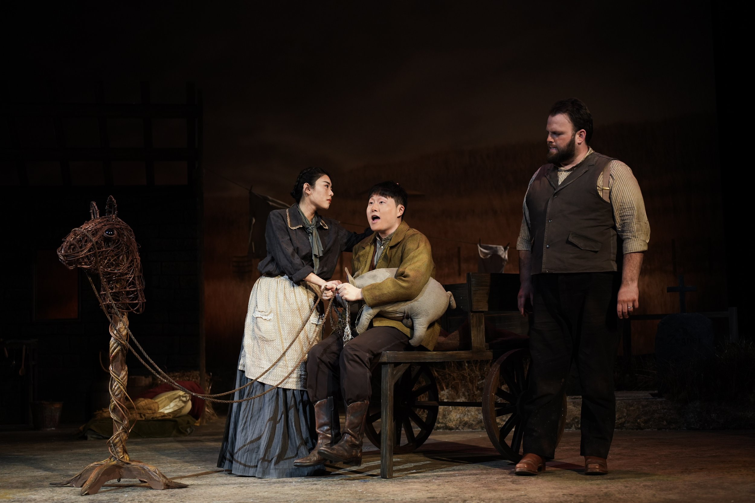 Theatre scene with three actors and a horse sculpture. A woman holds a man's hands, who appears distressed. Another man looks on, all dressed in period costumes, with a dark background.