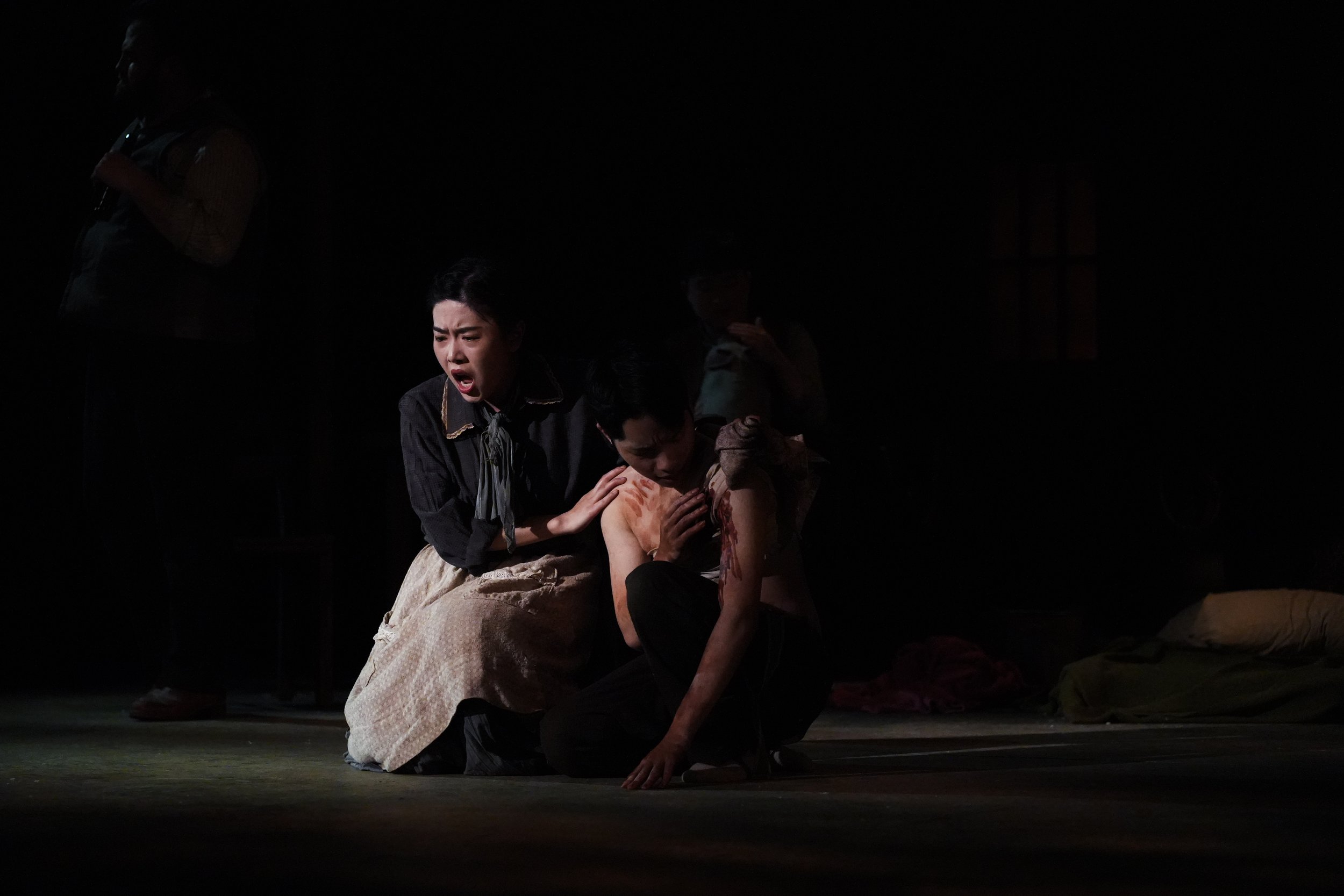 Theatrical stage scene with woman in distress, kneeling woman with blood on her arm, and coach in the dark.