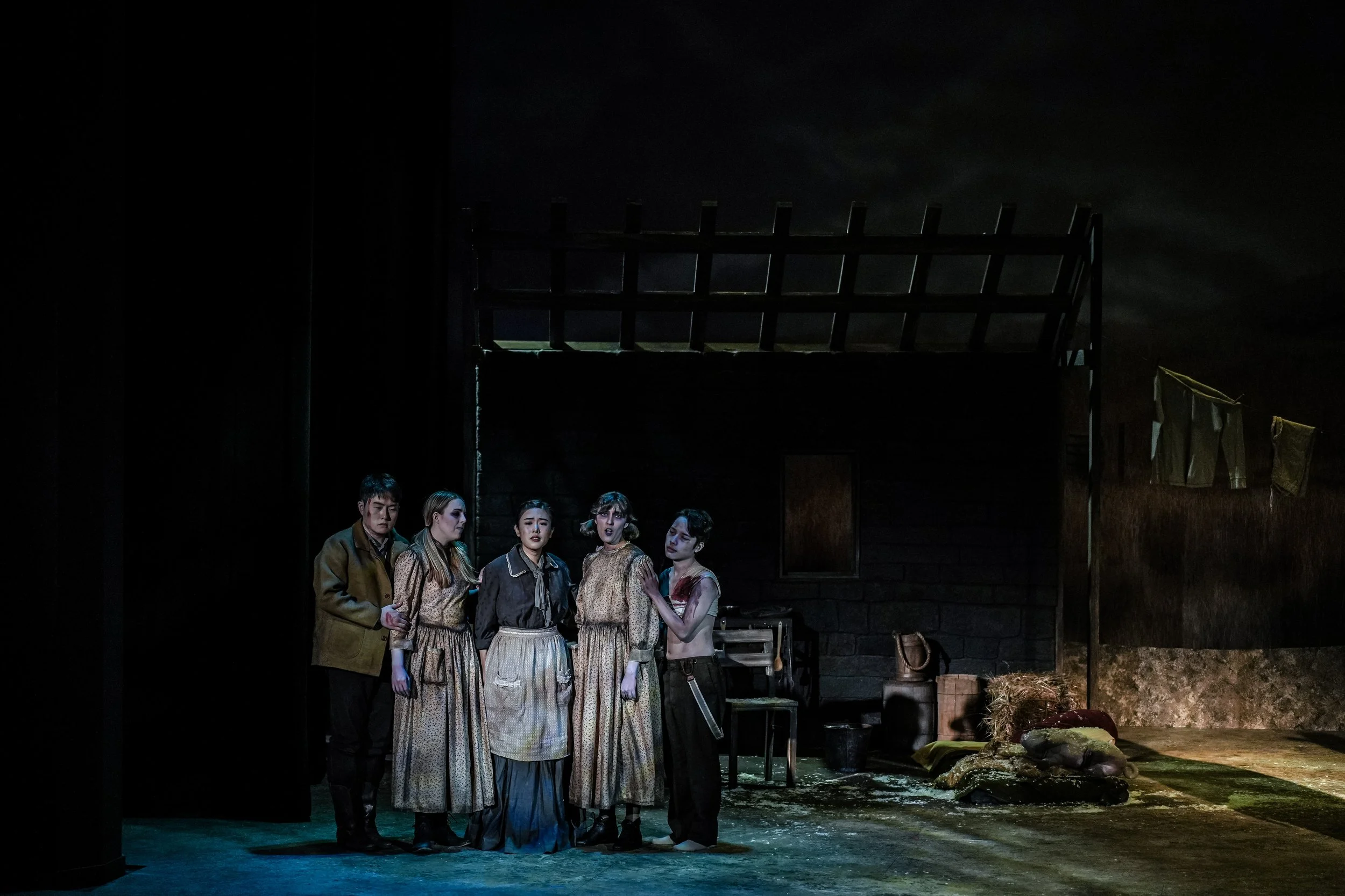 Group of six actors on stage during a theater performance, with simple set design including dark walls, a small window, and props like a chair, bag, and hay on the ground.