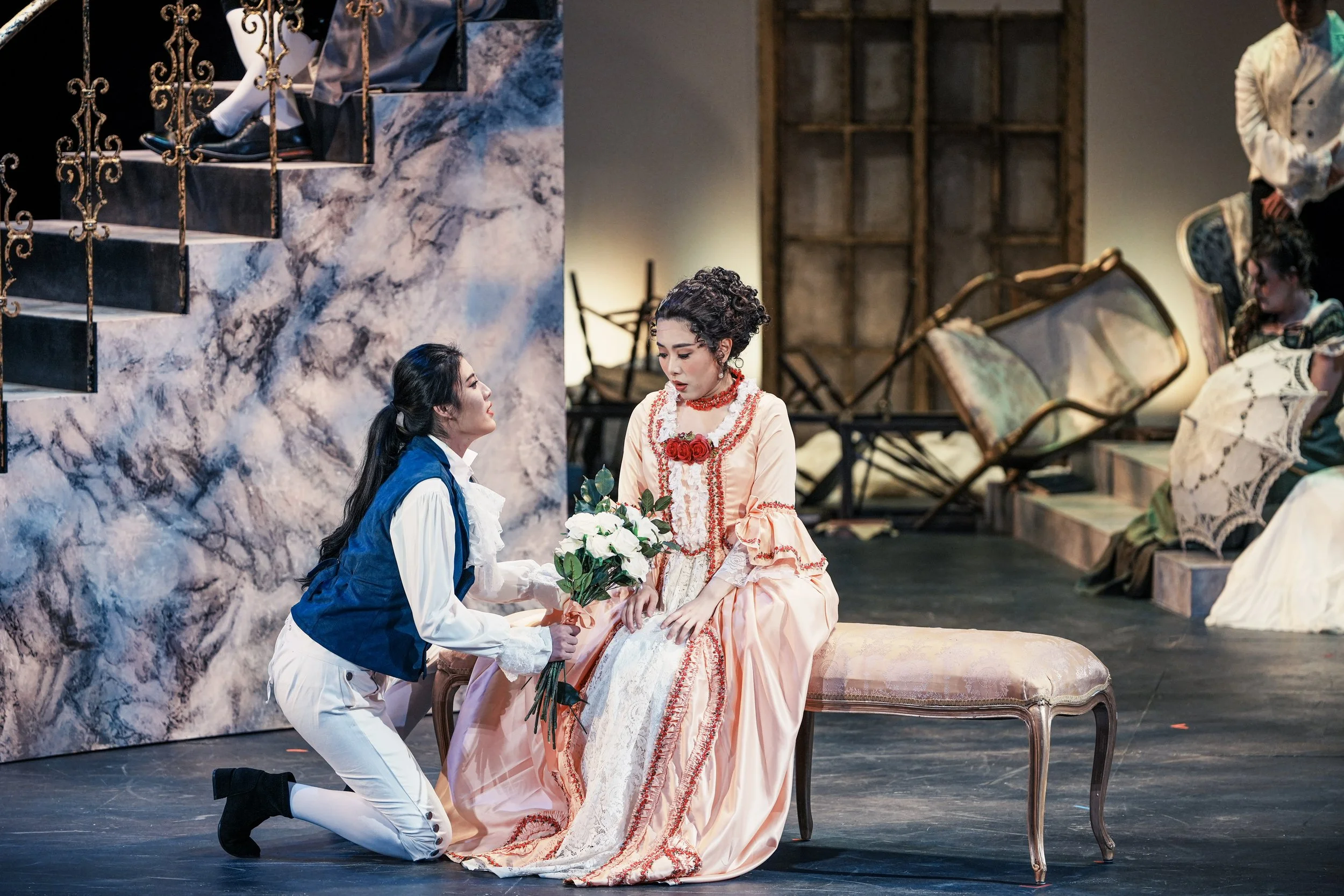 Two women on stage, one kneeling and offering flowers to the other seated on a bench, with a theatrical set designed like an elegant interior with stairs, chairs, and women in historical dresses in the background.