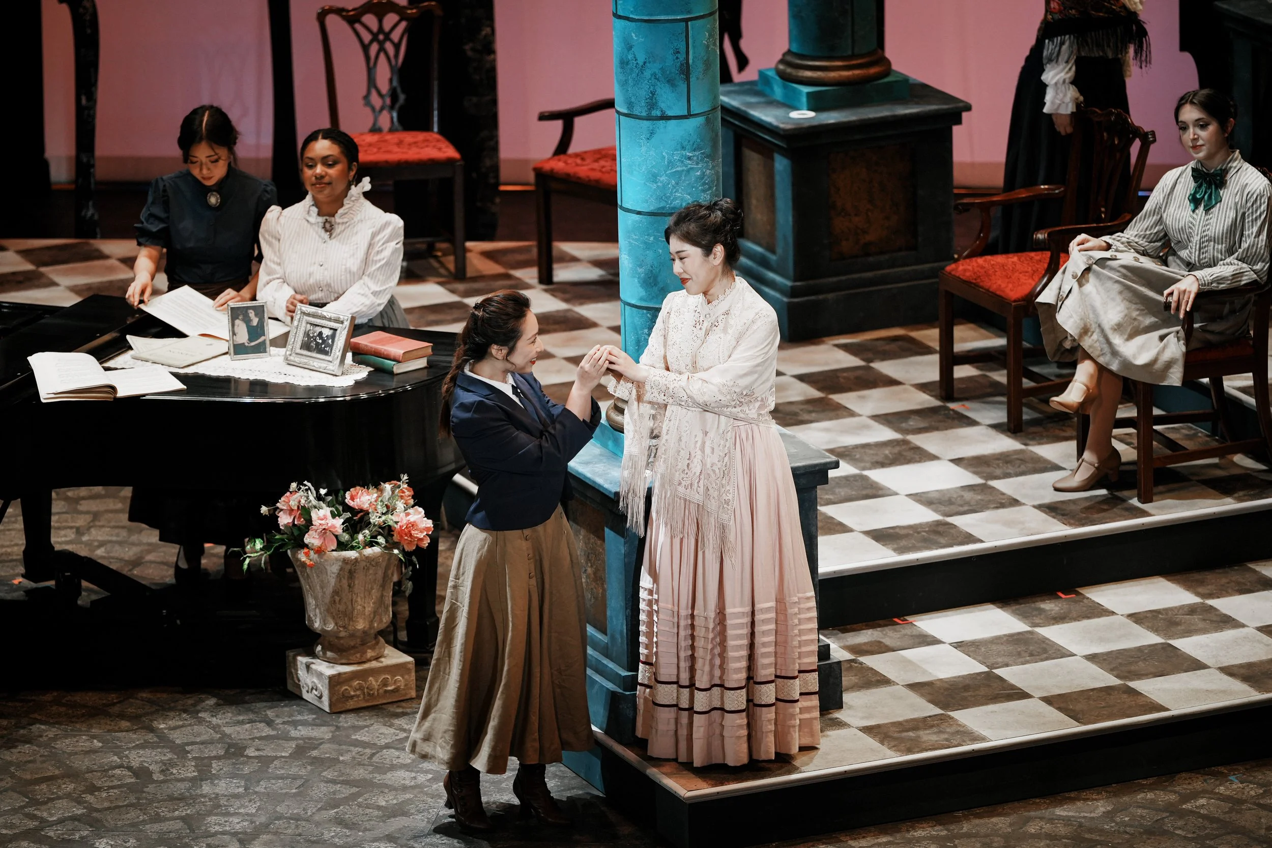 Stage scene with five women in period costumes, one woman on a bench, and a grand piano, set in a vintage-style room.
