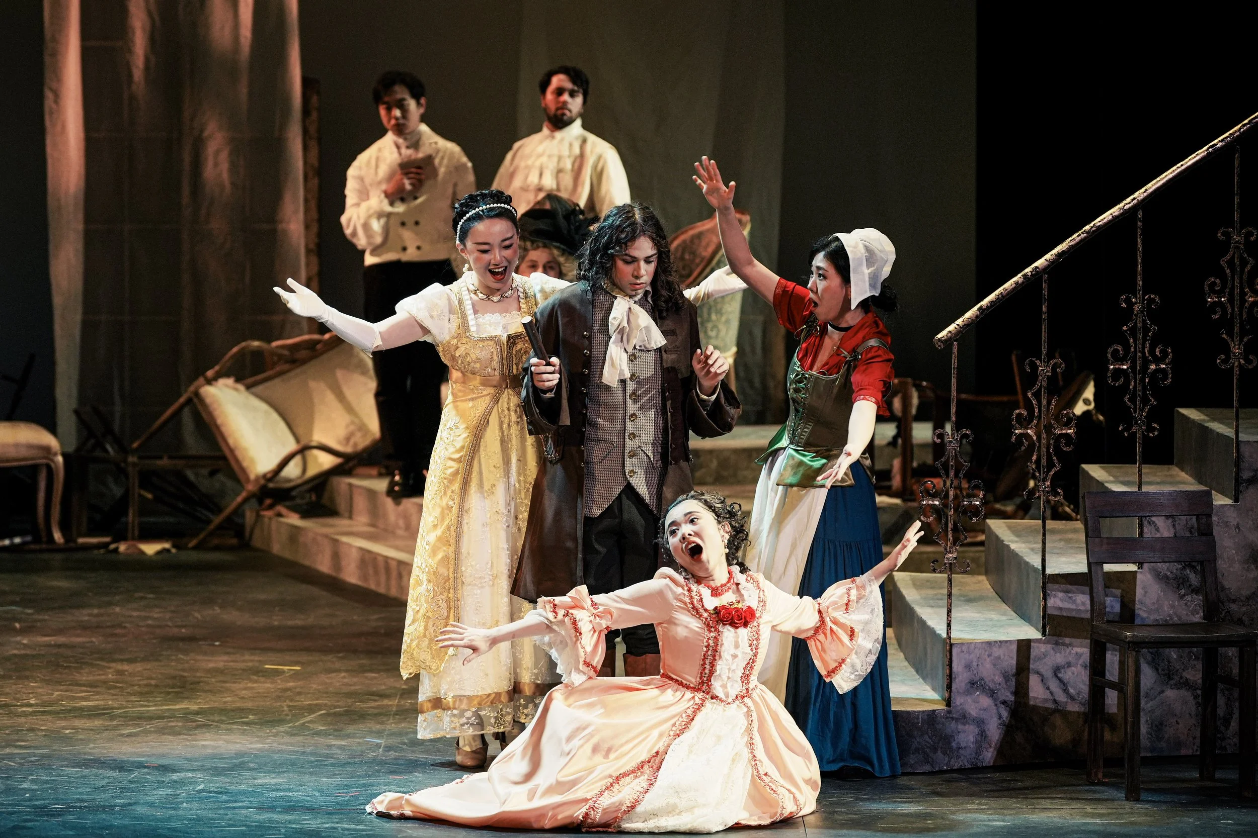 Actors in theatrical costume performing on stage, with one actress sitting on the floor with arms outstretched and others standing around, some gesturing and holding props, depicting a dramatic scene.