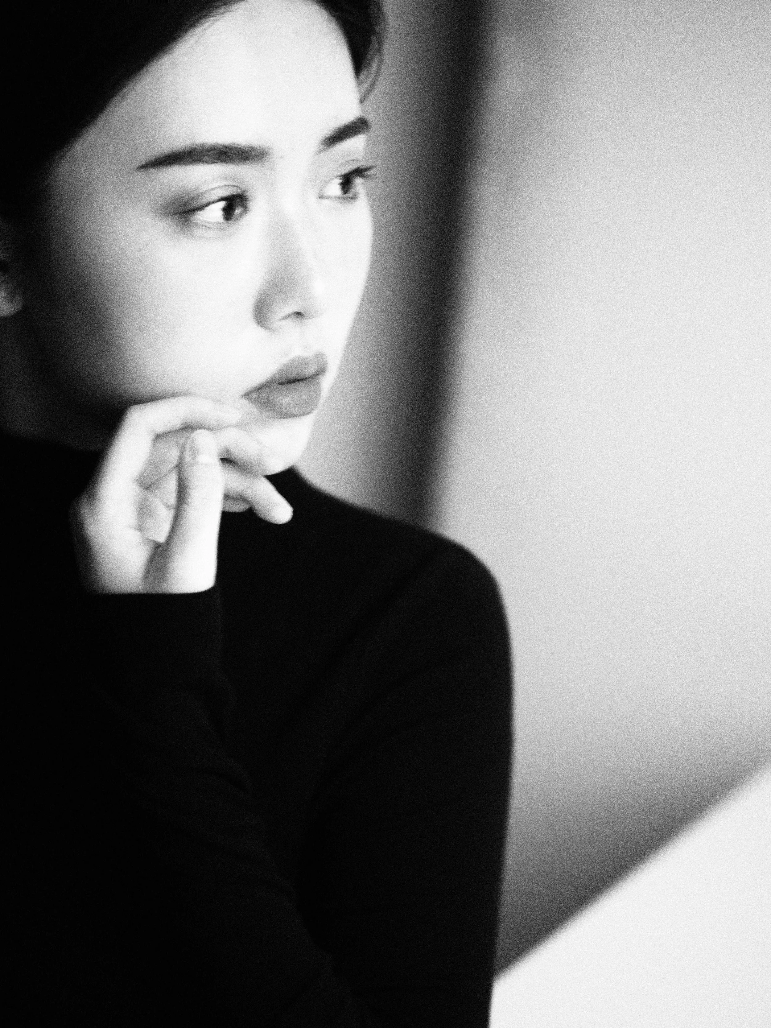 A black and white photograph of a woman with dark hair, wearing a dark top, looking thoughtfully to her left with her hand resting against her chin.