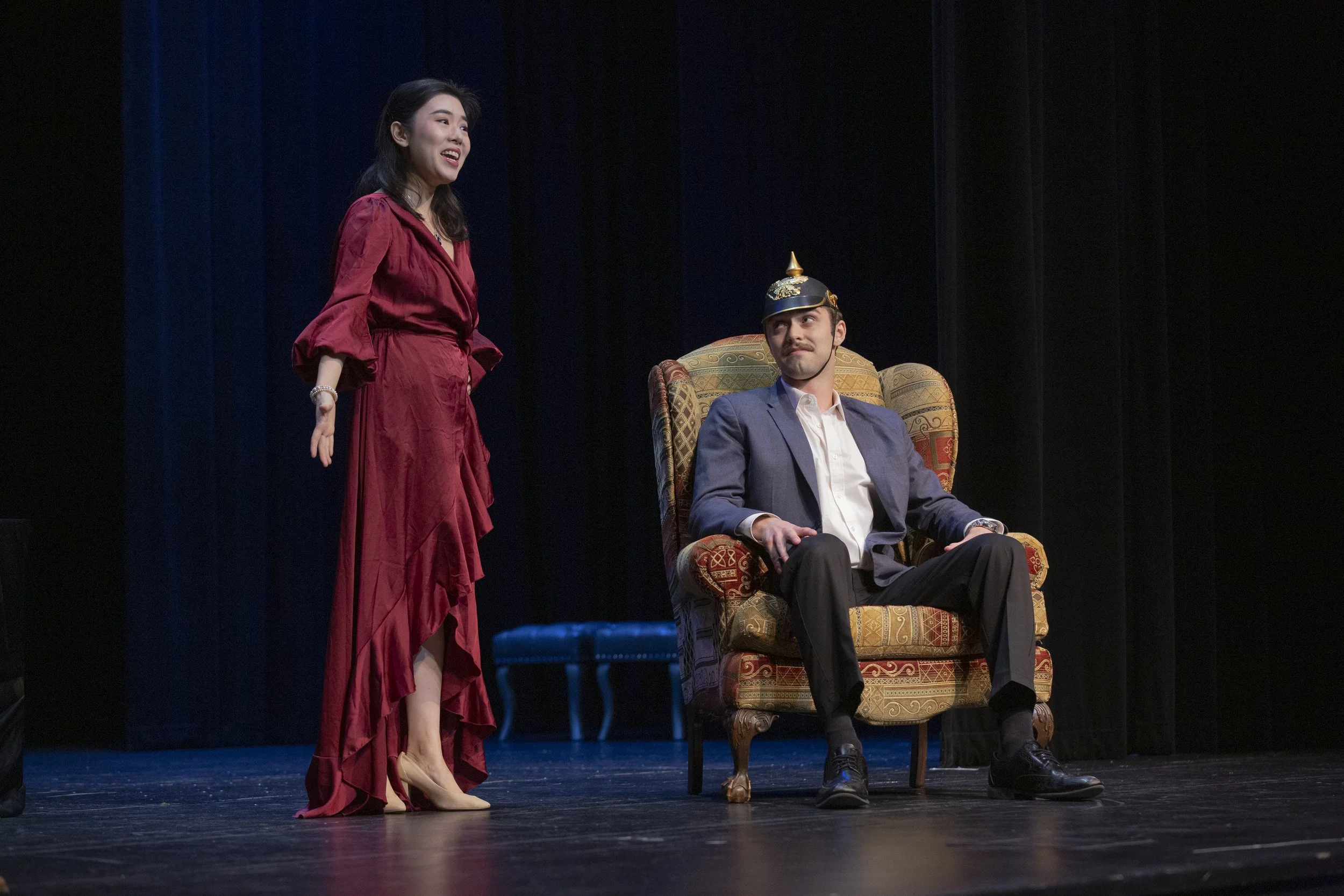 A woman in a red dress is standing and talking to a man in a suit, who is sitting in an ornate chair on stage. The man is wearing a conical hat and looks up at her.