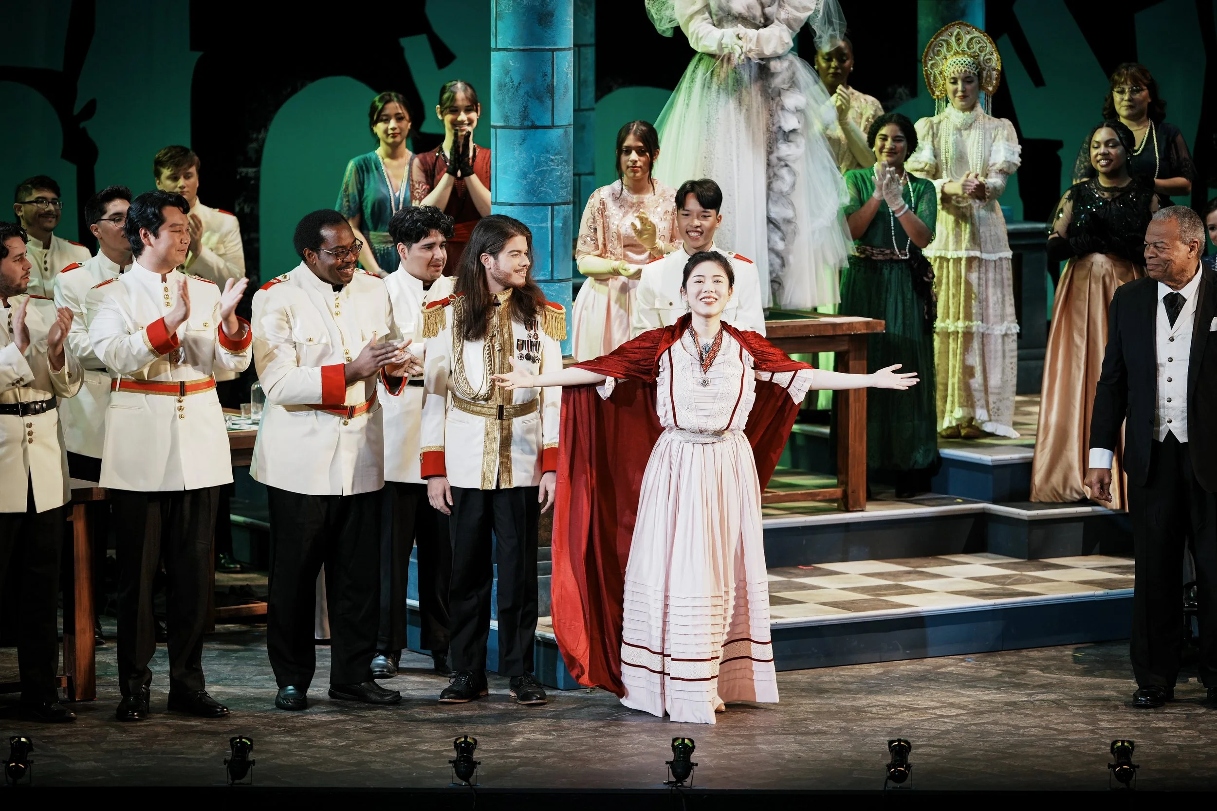 On stage, a woman dressed in a historical costume with outstretched arms stands in the center, surrounded by people in formal and traditional costumes, applauding in a theatrical setting.