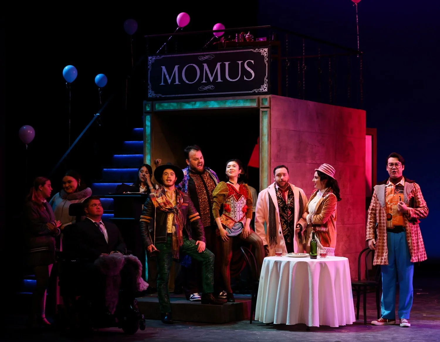 Theater stage scene with actors performing a play, with a sign reading 'MOMUS' above. Multiple actors are in colorful costumes, with some standing and others sitting, set around a table with drinks, in a decorated stage environment with stairs, ballo