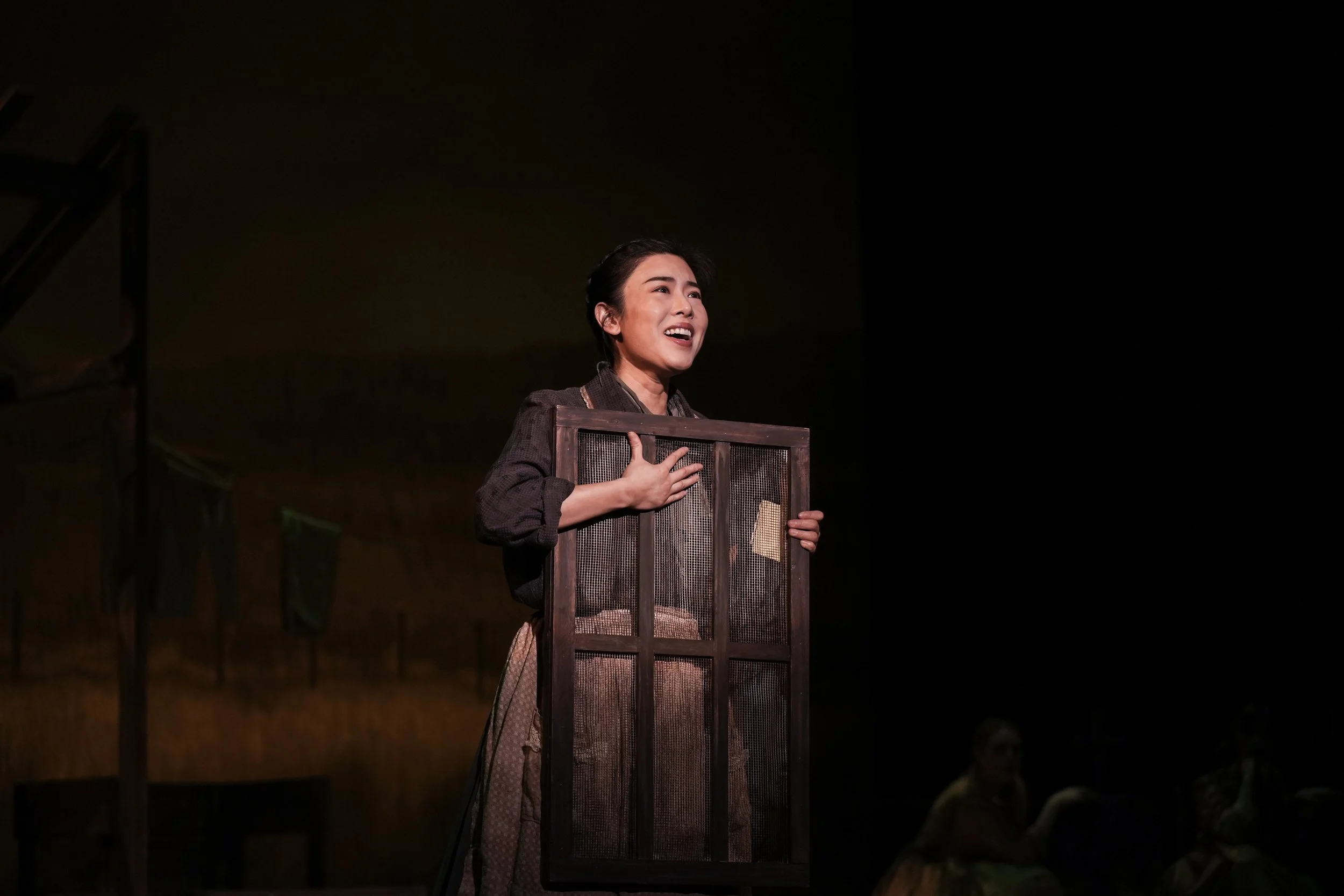 A woman on stage holding a screen or window frame, singing or speaking with emotion, while another person in the background is seated and partially visible.