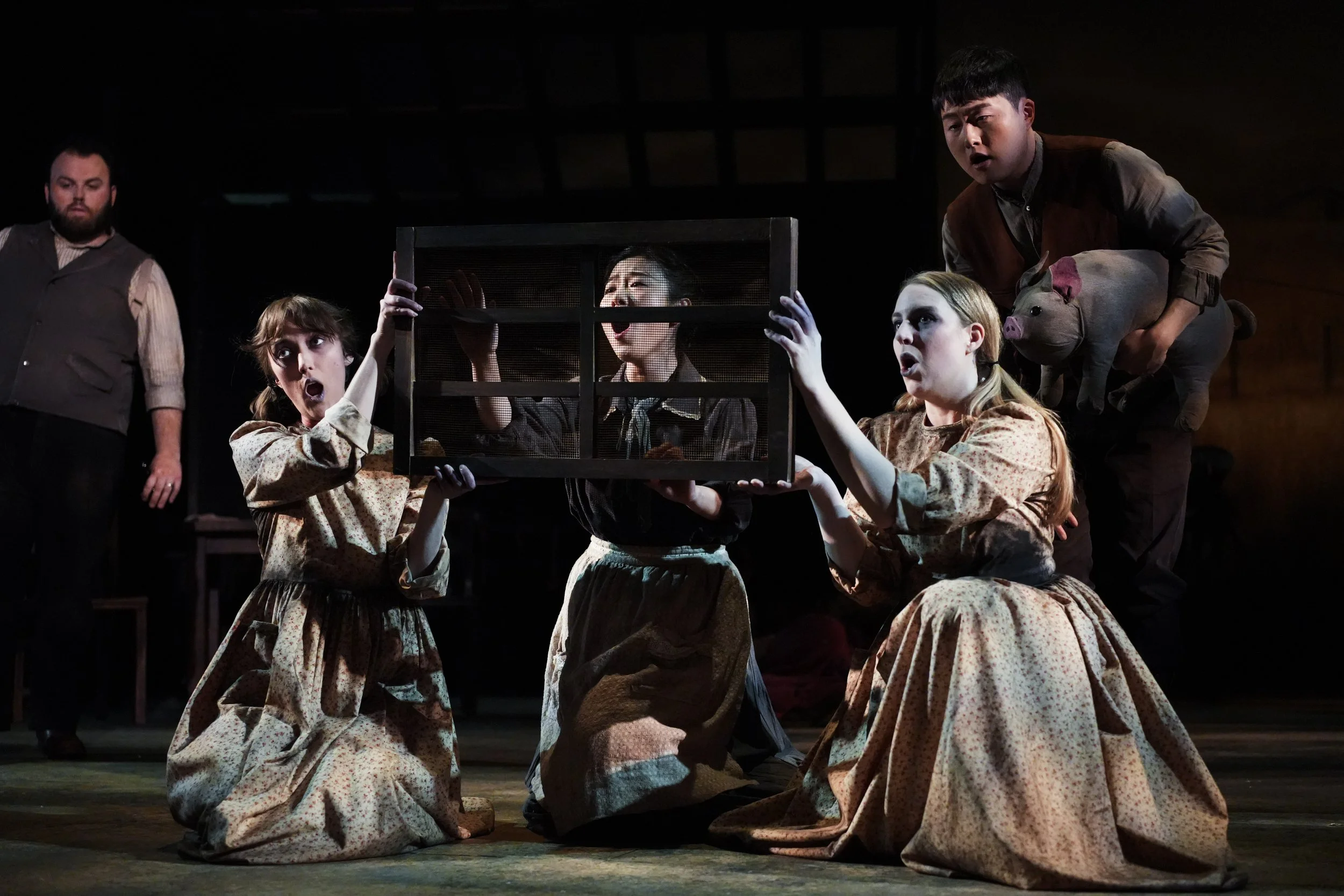 Theatrical scene with children in period costumes, two kneeling and holding a cage, others standing with a pig puppet, all expressive as if in a play.